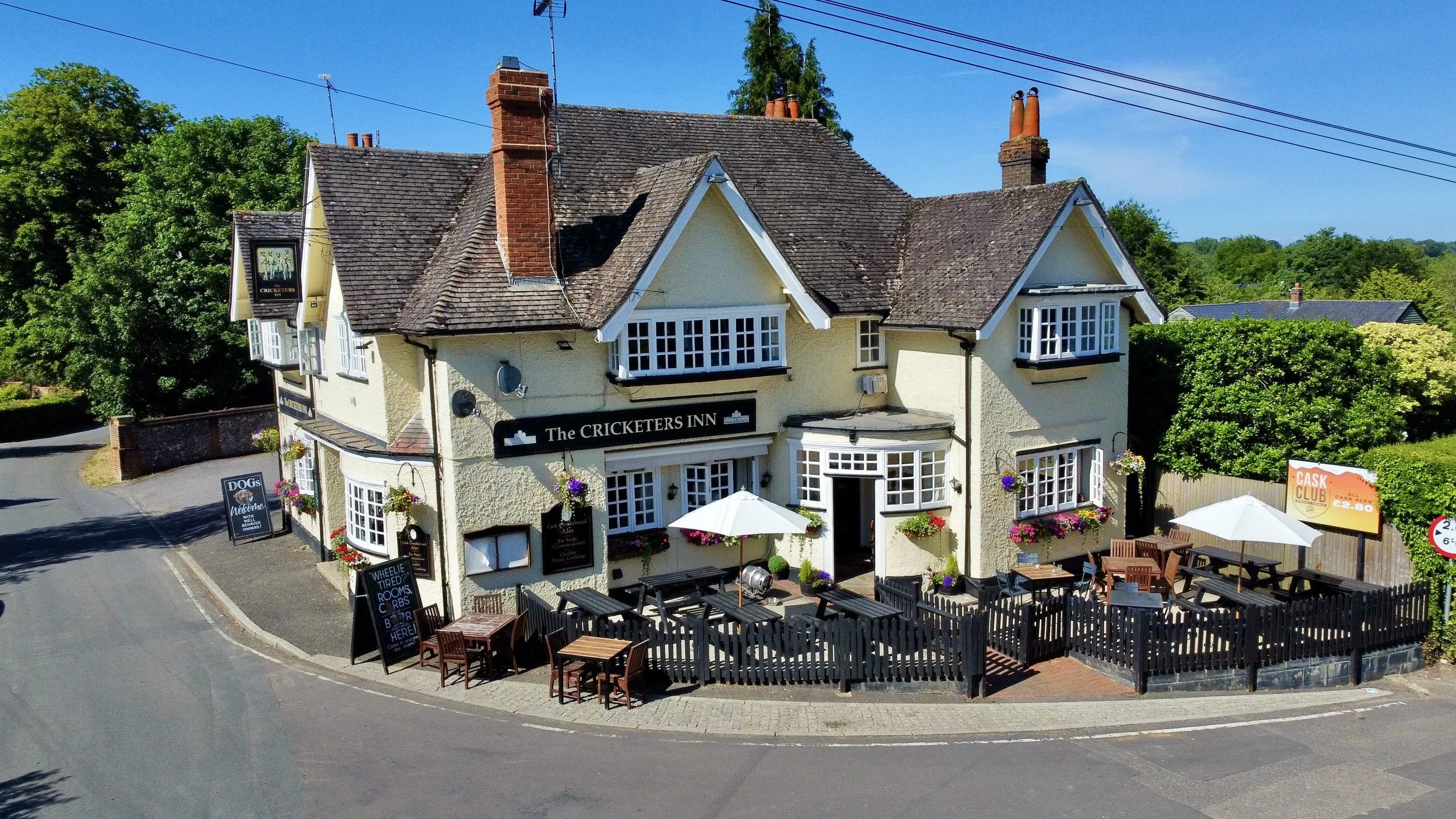 An aerial view of The Cricketers Inn, a two-story pub with outdoor seating, flower boxes, and umbrellas, located on a curved street with greenery and trees in the background.