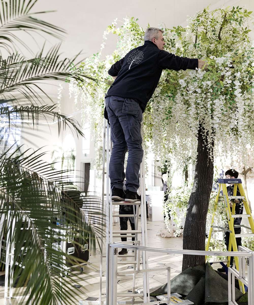 A man standing on a ladder arranging white flowers and greenery on an indoor artificial tree for decoration.