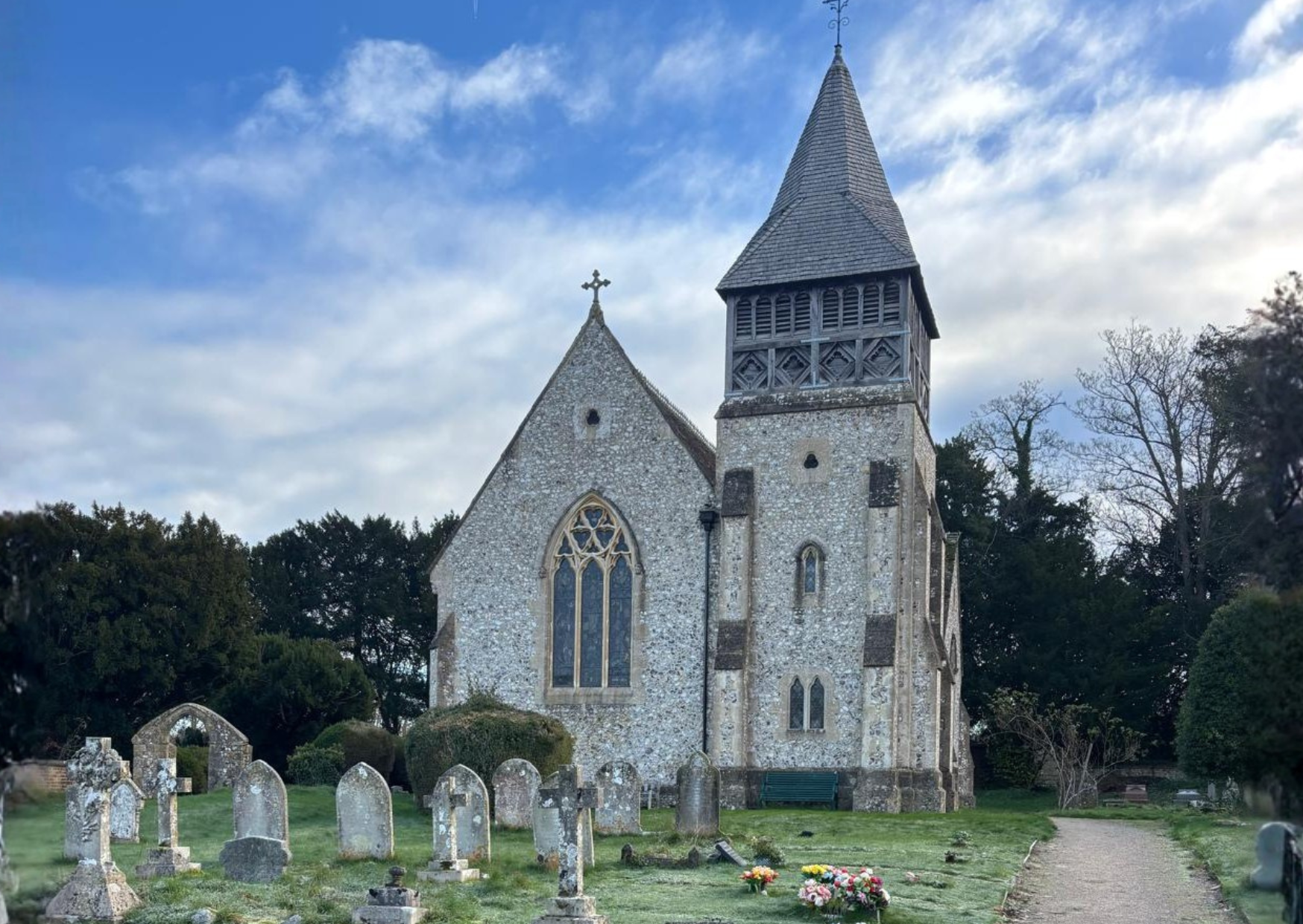 An old stone church with a pointed steeple and stained glass windows, set against a partly cloudy sky, with a graveyard in the foreground.