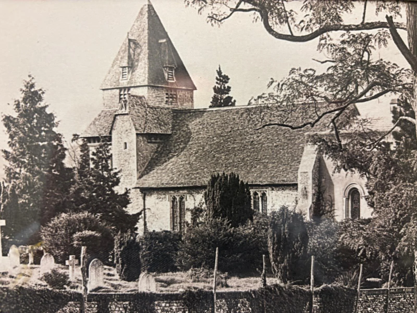 An old black and white photo of a church surrounded by trees, with a prominent tower and pointed roof, and a graveyard in the foreground.