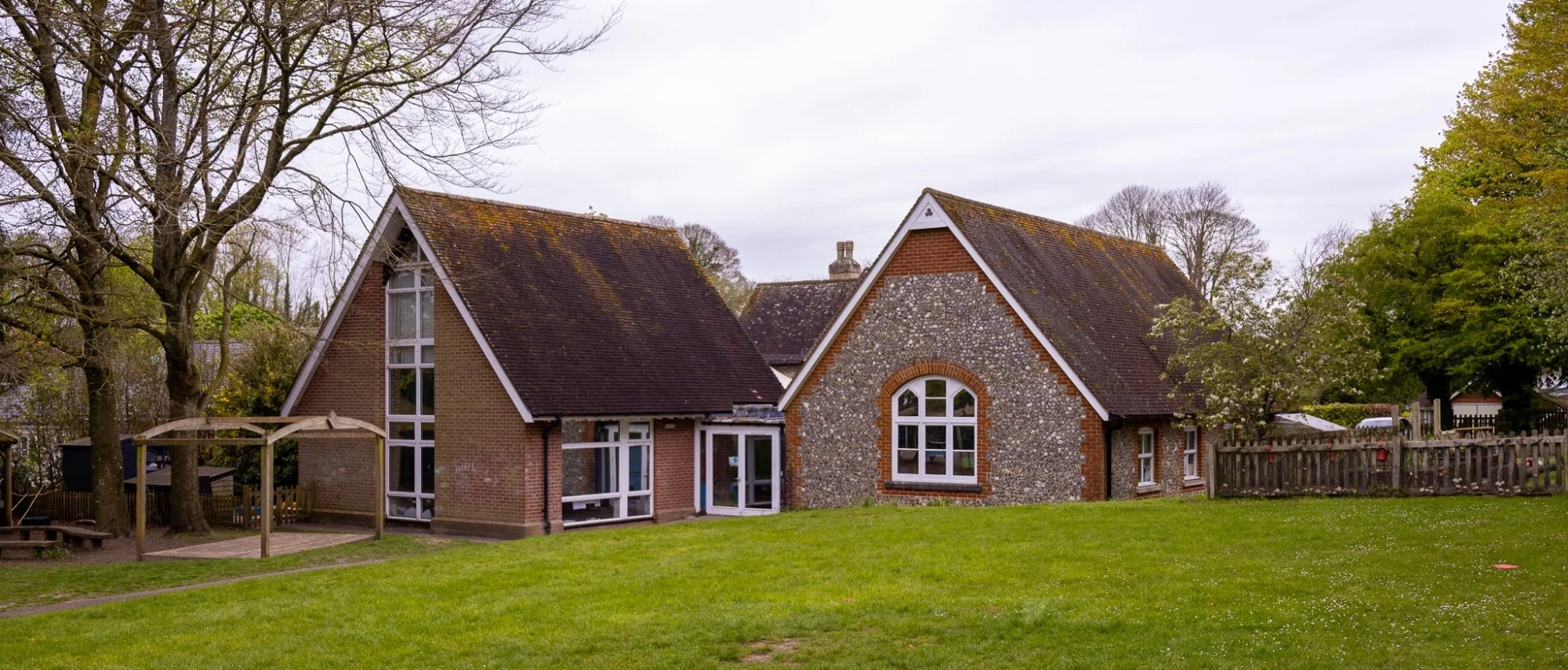 A large brick house with a sloped roof, windows, and a small enclosed front yard with a grassy lawn, trees, and a wooden fence on a cloudy day.
