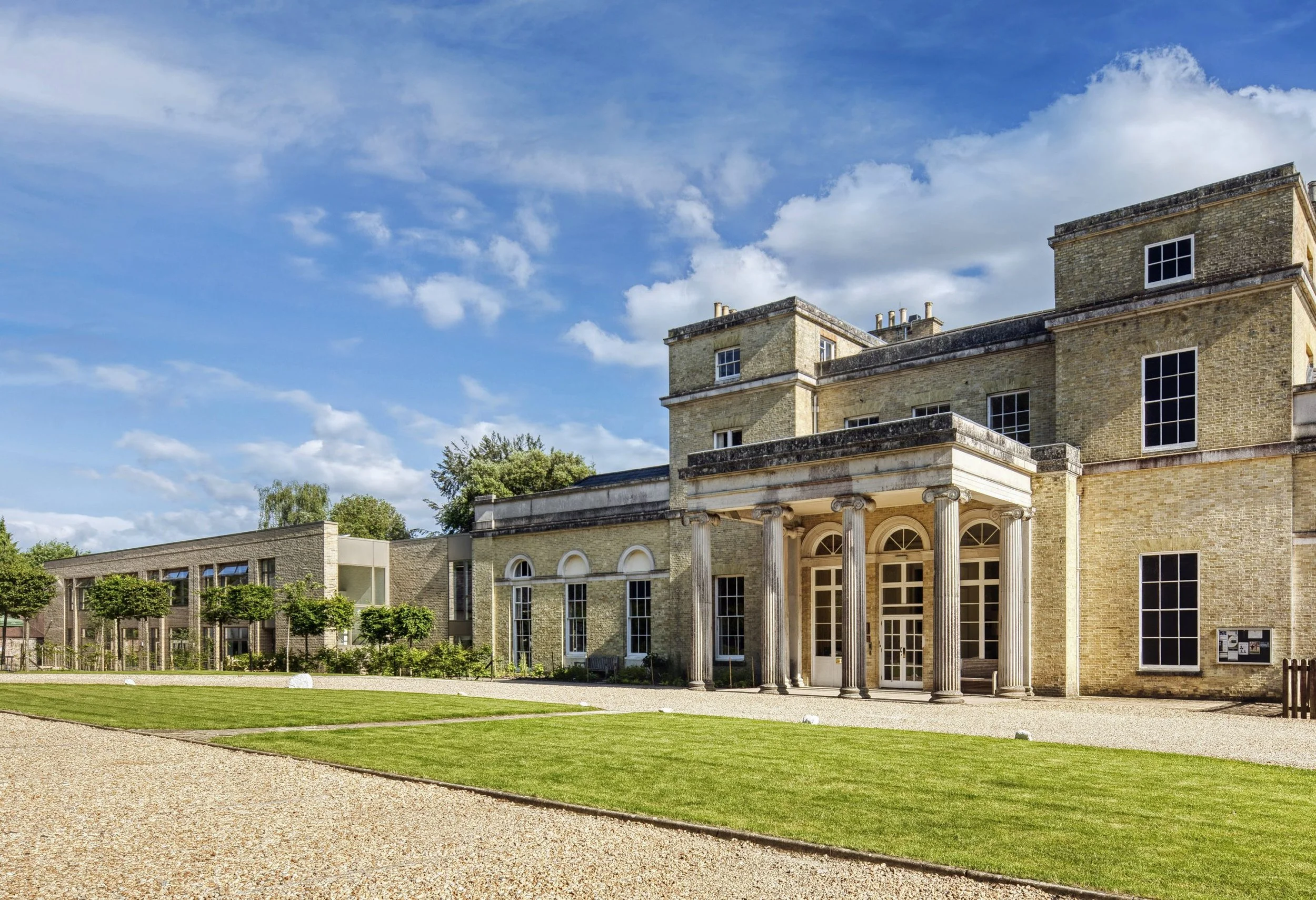 A large, historic brick building with columns at the entrance, multiple windows, surrounded by a well-maintained lawn and gravel paths, under a partly cloudy sky.