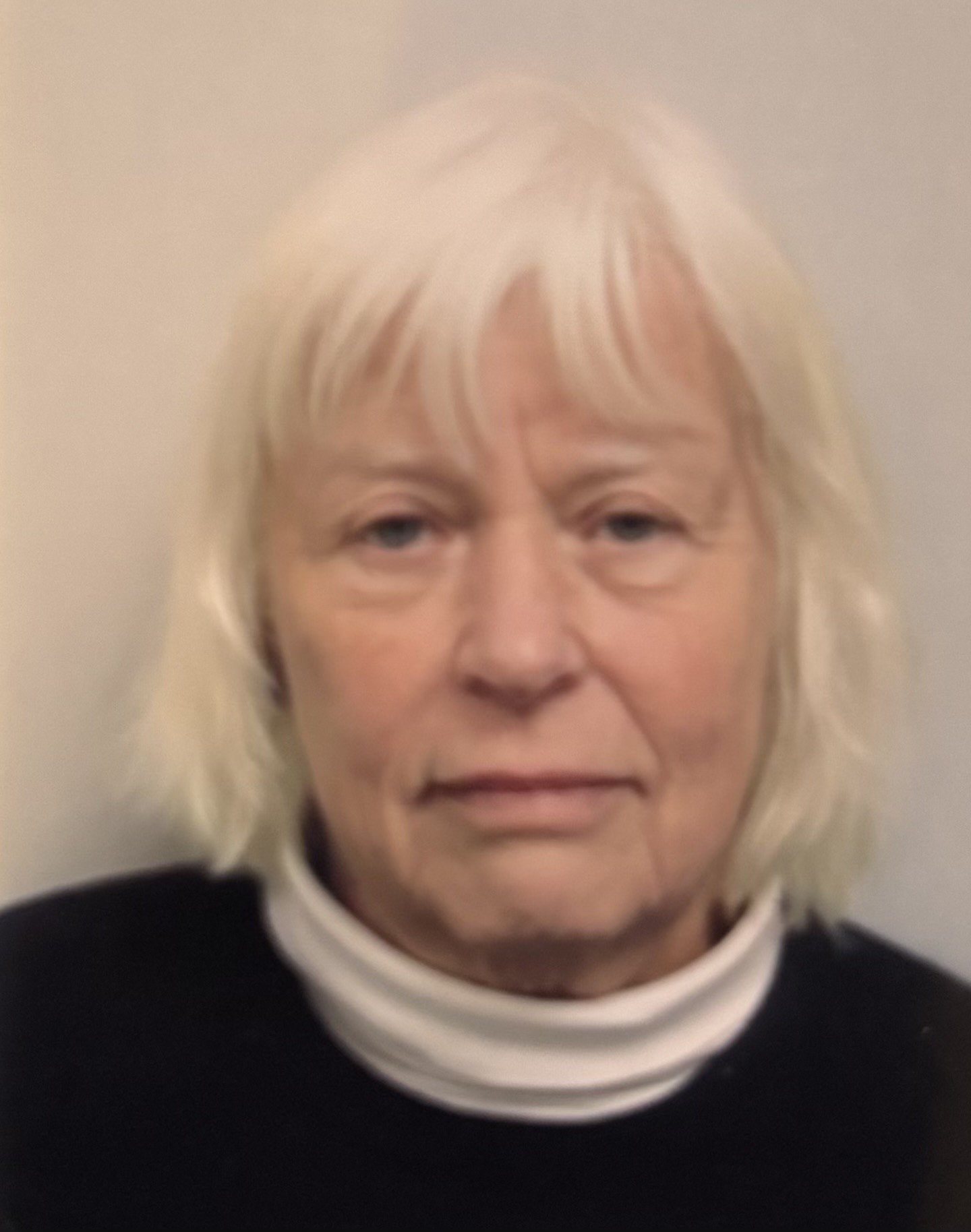 Close-up photo of an older woman with shoulder-length white hair, light skin, wearing a black top and white turtleneck, against a plain light-colored background.