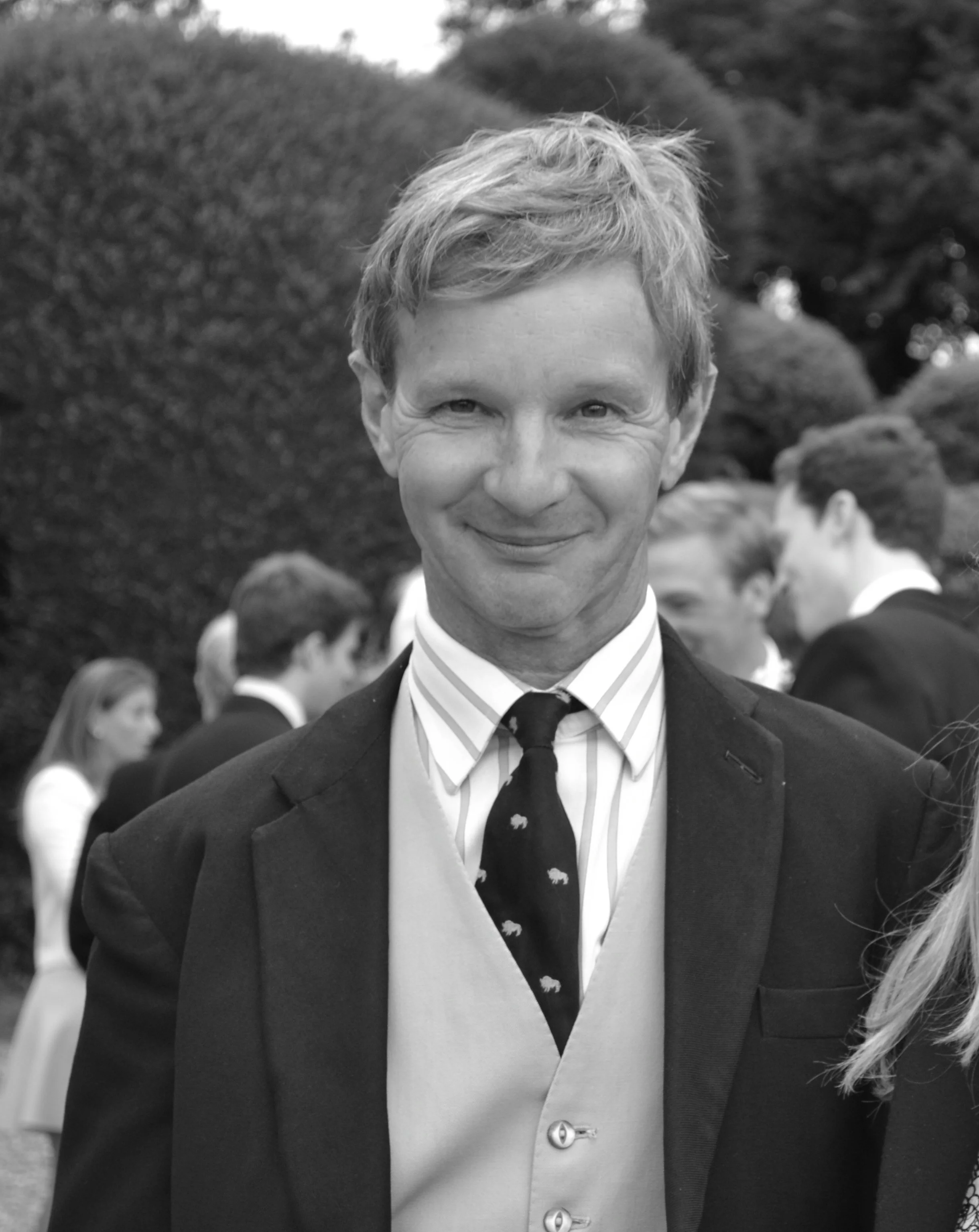 A smiling man in formal attire at an outdoor event, with a group of people in the background.