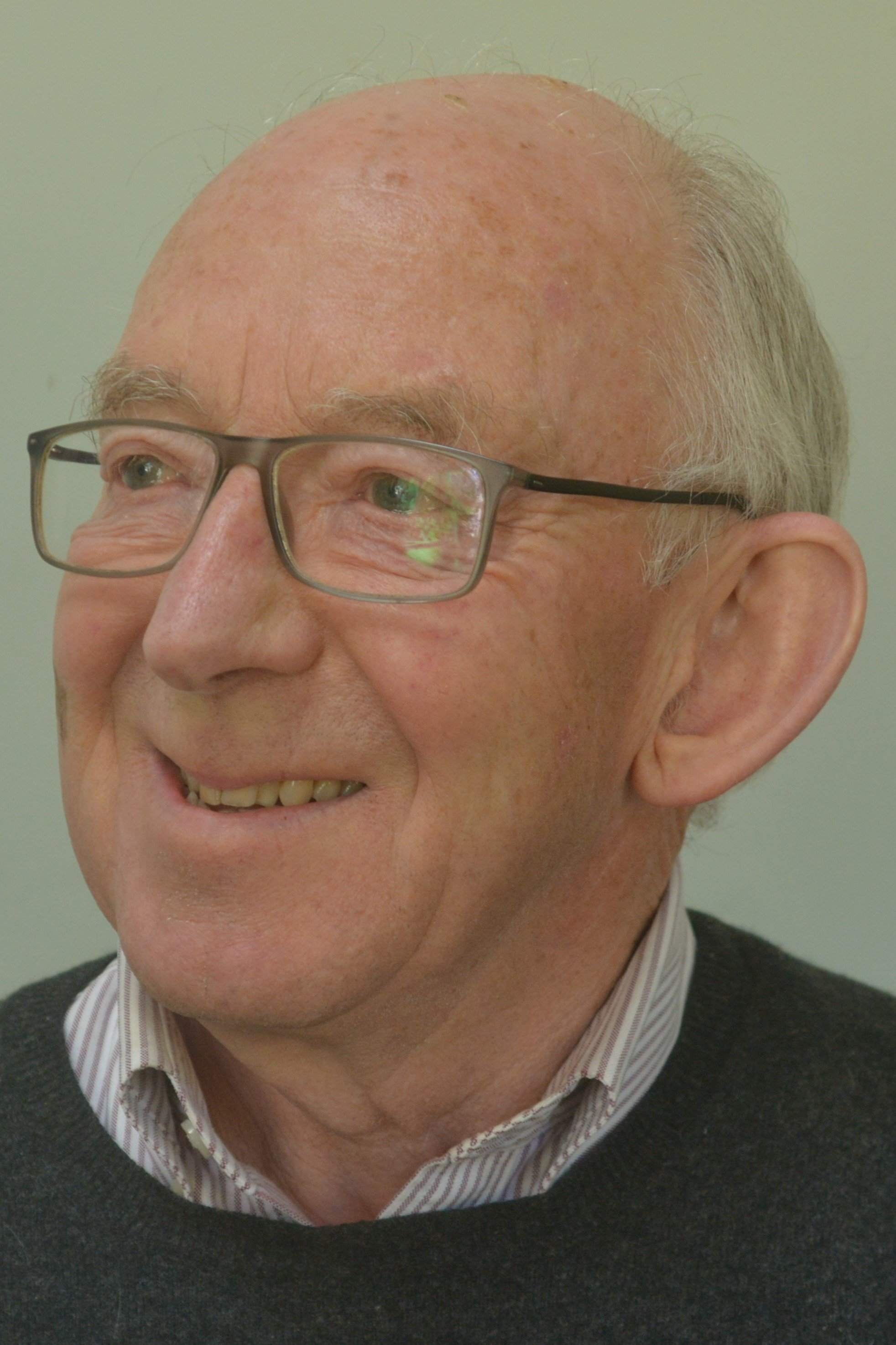 Close-up of an elderly man smiling, wearing glasses, a gray sweater, and a collared shirt, with a plain background.