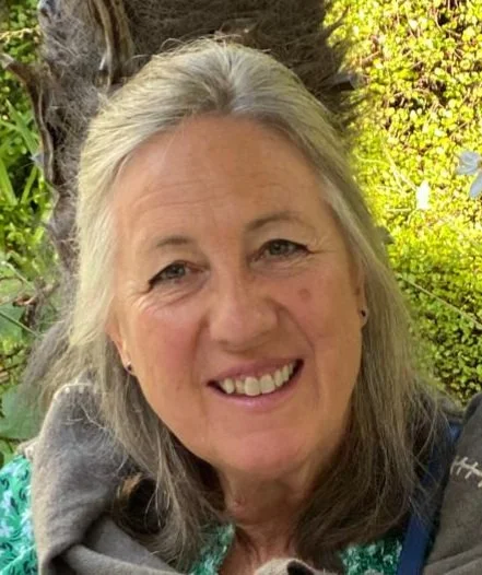 A middle-aged woman with long gray hair smiling outdoors among trees and green foliage.