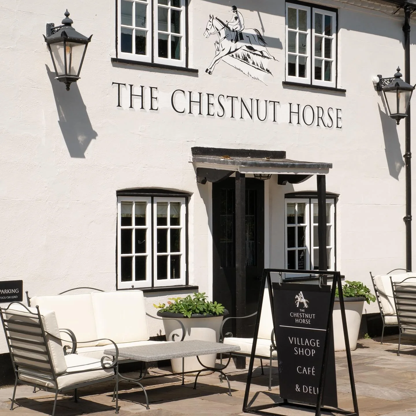The exterior of The Chestnut Horse pub with outdoor seating, large white planters, a black chalkboard sign, and two black wall-mounted lamps.