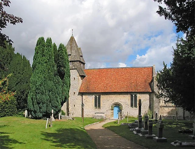 A historic stone church with a red-tiled roof, surrounded by trees and a graveyard, under a partly cloudy sky.