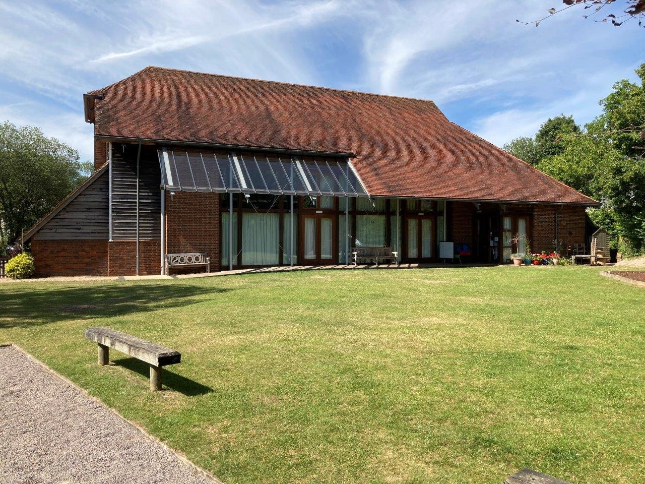 A large brick house with a sloped red roof and glass doors on the ground level. There are outdoor benches, potted plants, and a small yard with green grass under a partly cloudy sky.