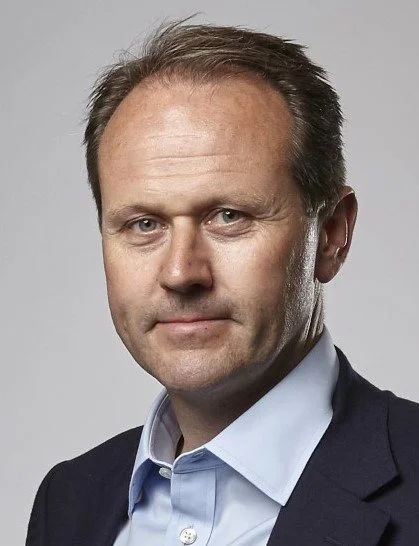 Close-up portrait of a middle-aged man with short brown hair, wearing a light blue dress shirt and a dark suit jacket, against a gray background.