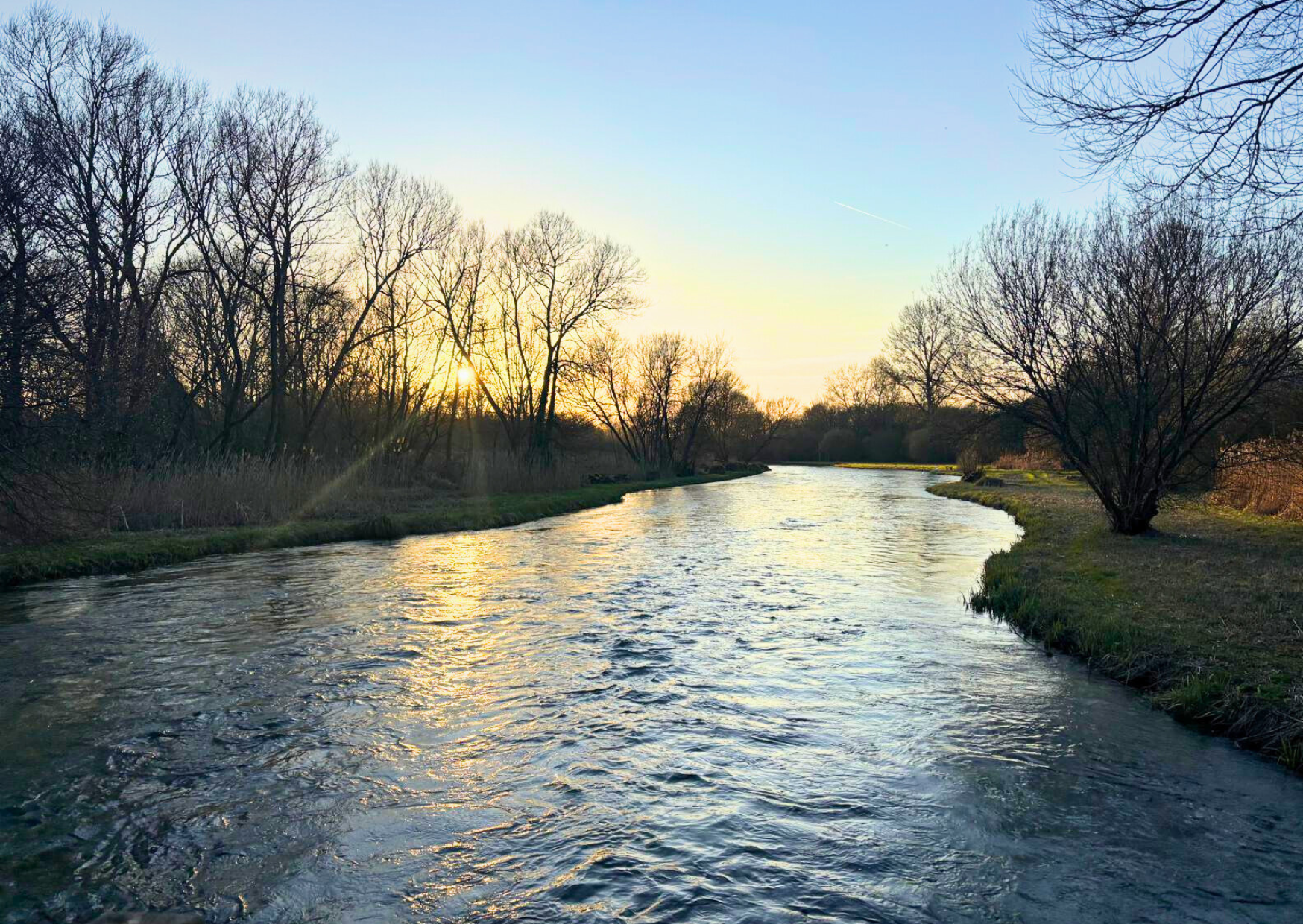 A serene river curves through a landscape during sunset, with bare trees on both sides and a clear sky with a faint star or planet visible.