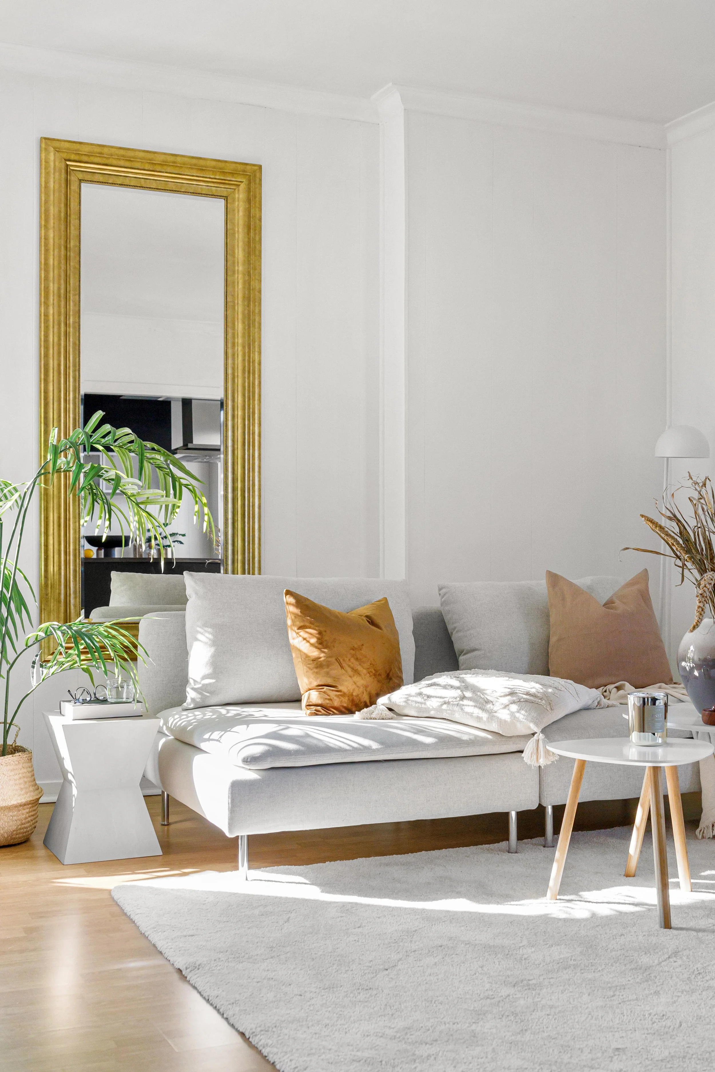 Salon moderne avec canapé gris clair, coussins en velours doré et beige, tapis blanc, plantes vertes, miroir encadré en or, lampadaire blanc, table basse ronde blanche avec bougies, mur blanc, décoration minimaliste et lumineuse.