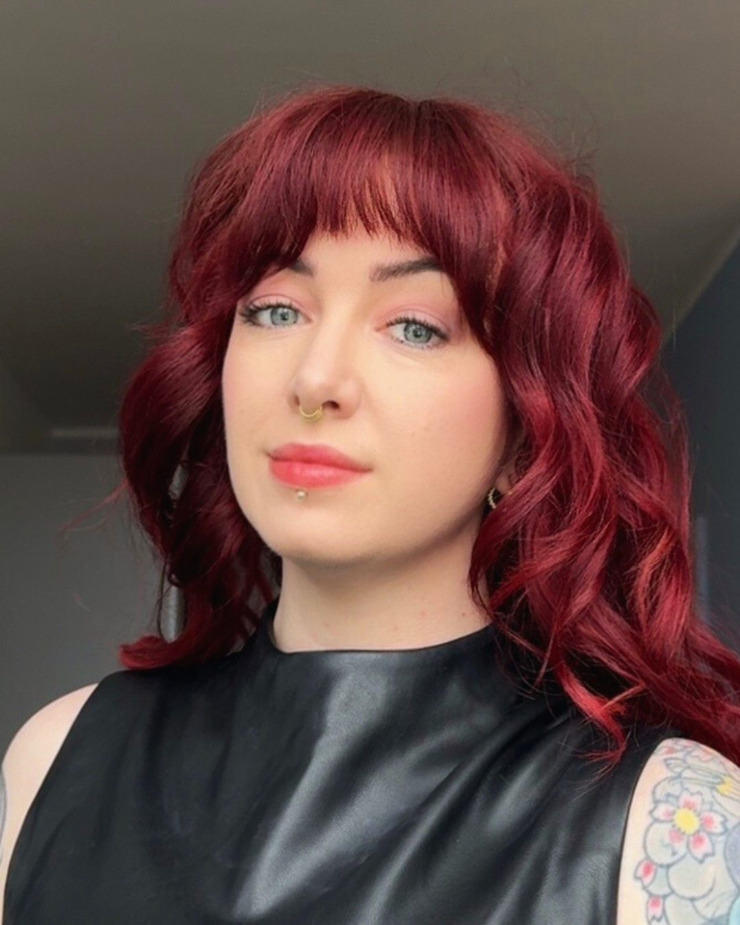 Portrait of a woman with red curly hair, blue eyes, and a septum piercing; wearing a black sleeveless top and earrings, showing a tattoo on her upper arm.