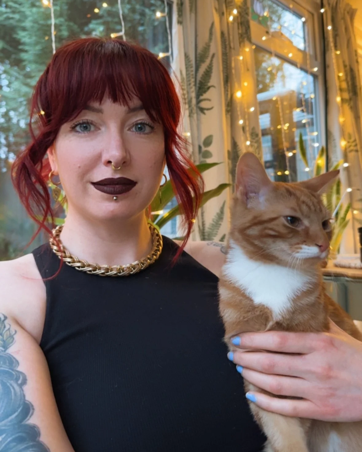 A woman with red hair and dark lipstick holding an orange and white tabby cat in a room decorated with string lights and plants.
