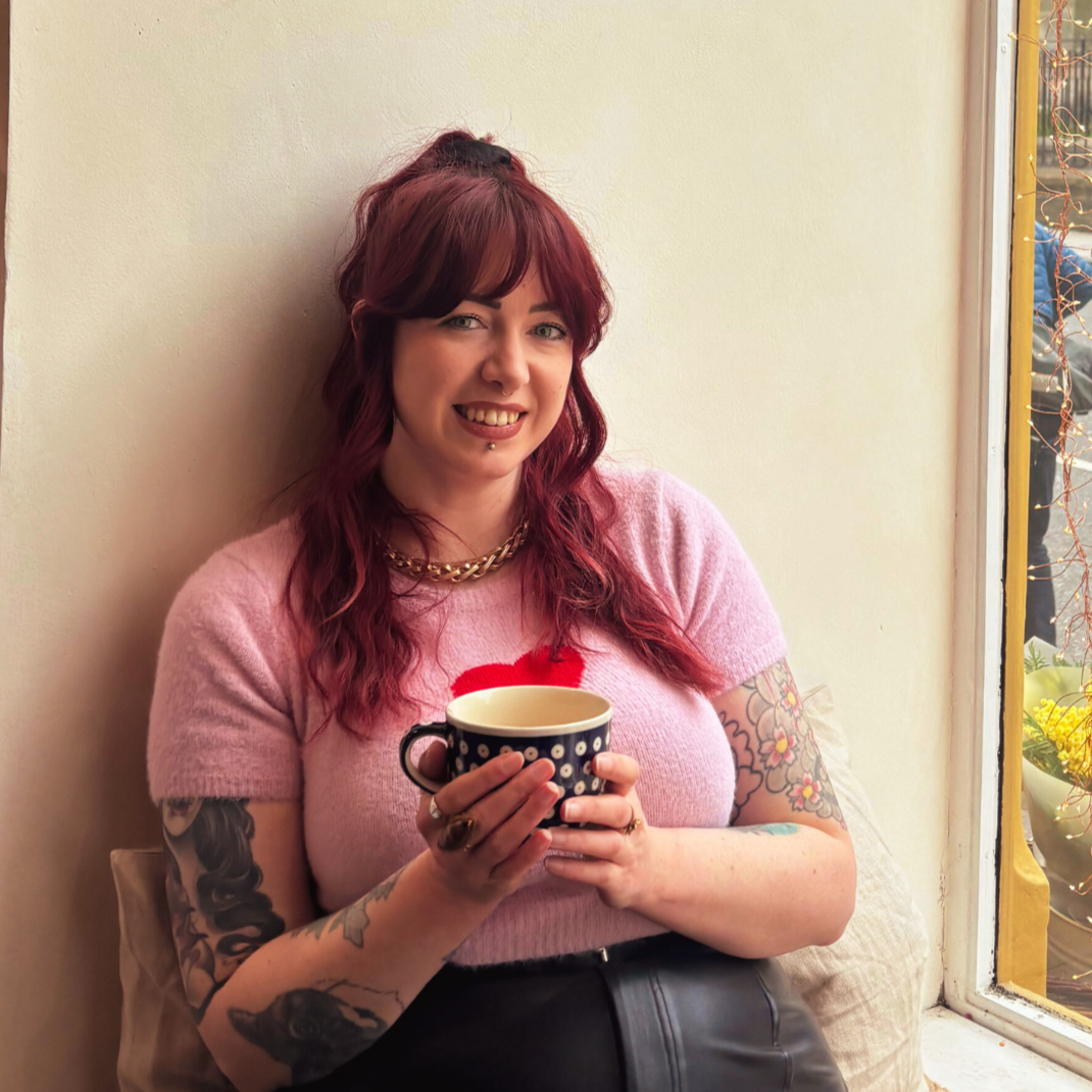 A woman with wavy red hair tattoos and pierced lips wearing a black top, taking a photo in a room with framed gaming artwork on the wall behind her.