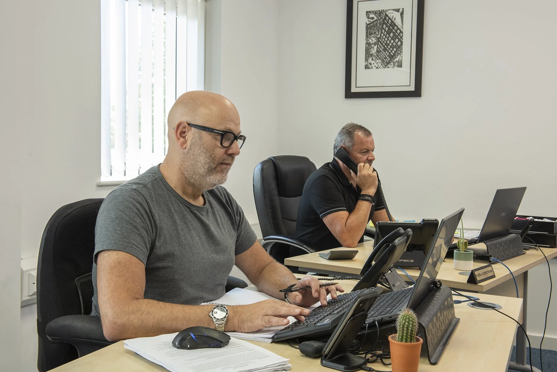 Two men working at desks. one of them is on phone. 