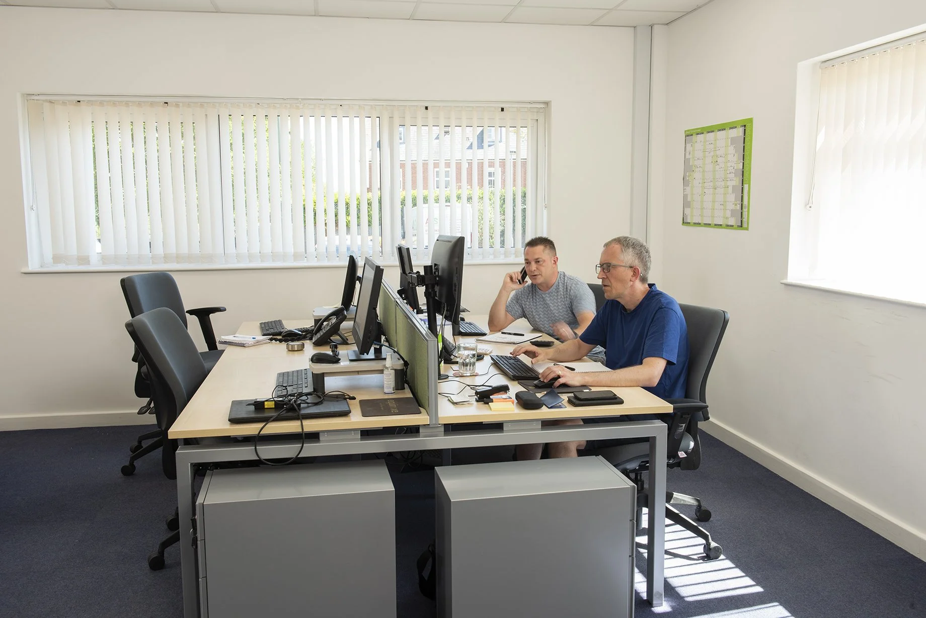 “Two people seated at a shared desk setup with multiple monitors, working in a bright office with large windows and blinds.”