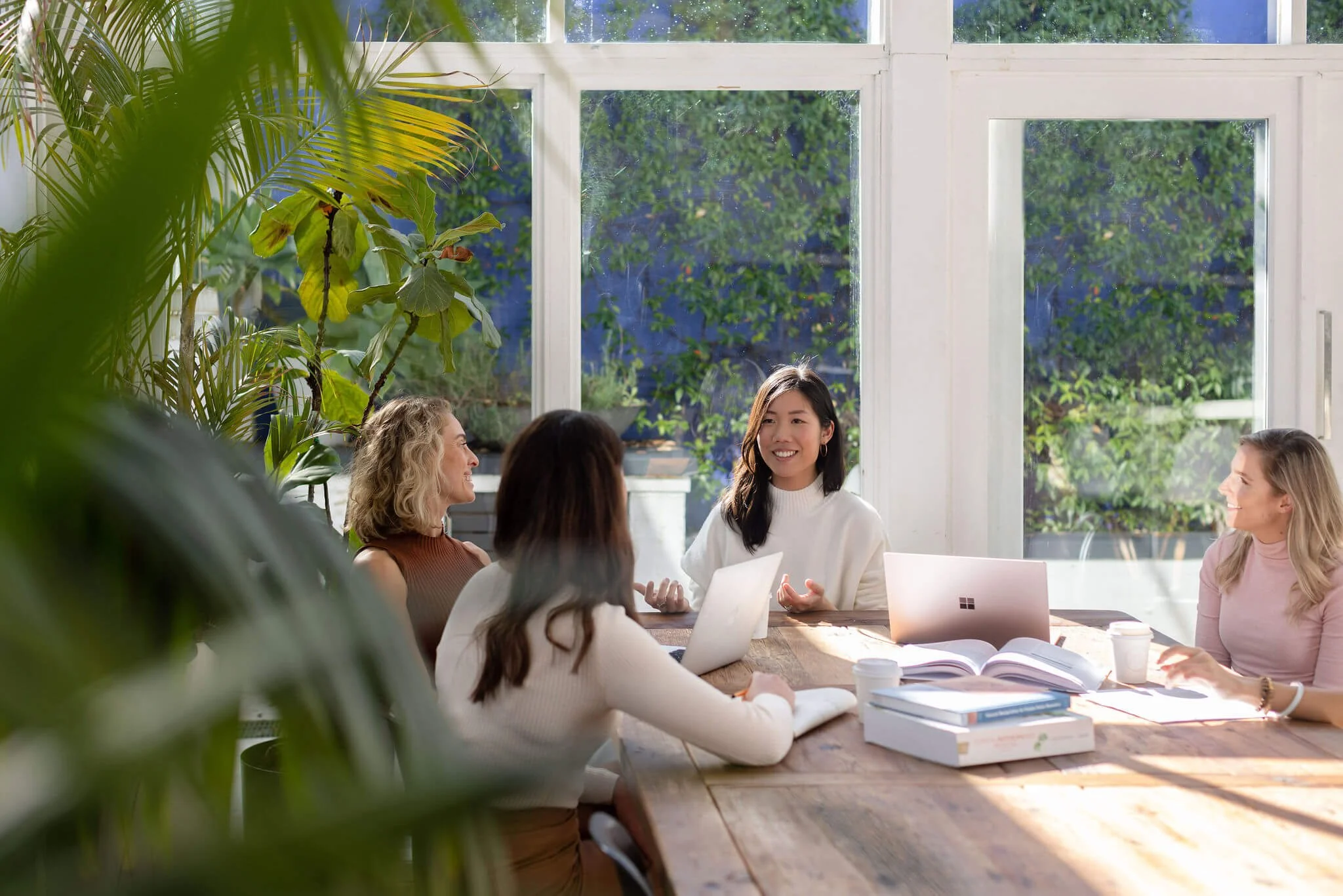 Group of five women sitting around a wooden table in a bright room with large windows and greenery outside, engaged in conversation with laptops and books in front of them.