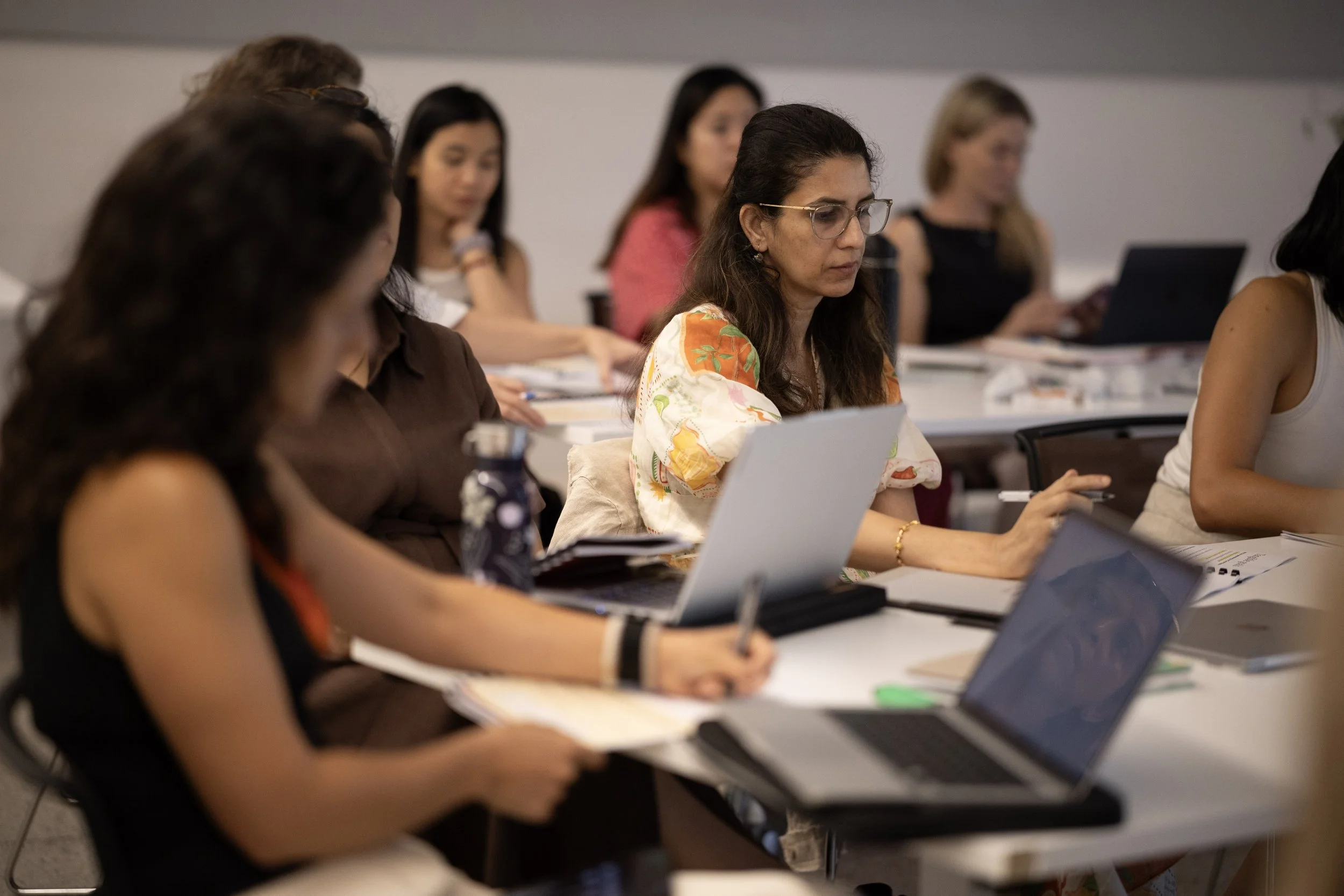 Group of women attending a seminar or conference, sitting at tables with laptops and papers, focusing on the presentation or speaker.