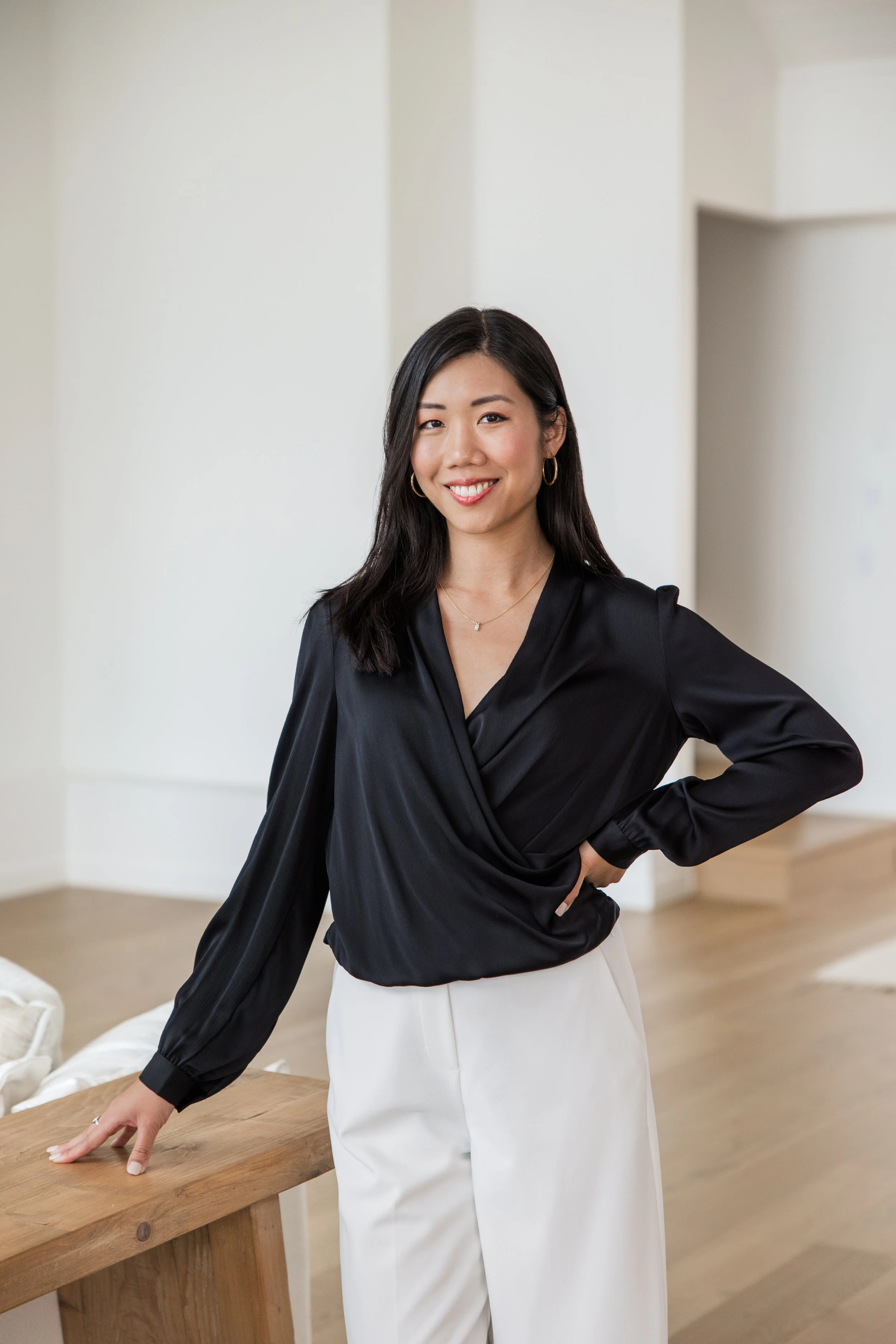 A woman wearing a black satin wrap blouse and white high-waisted trousers, smiling and standing in a bright, modern room with white walls and wooden floors.