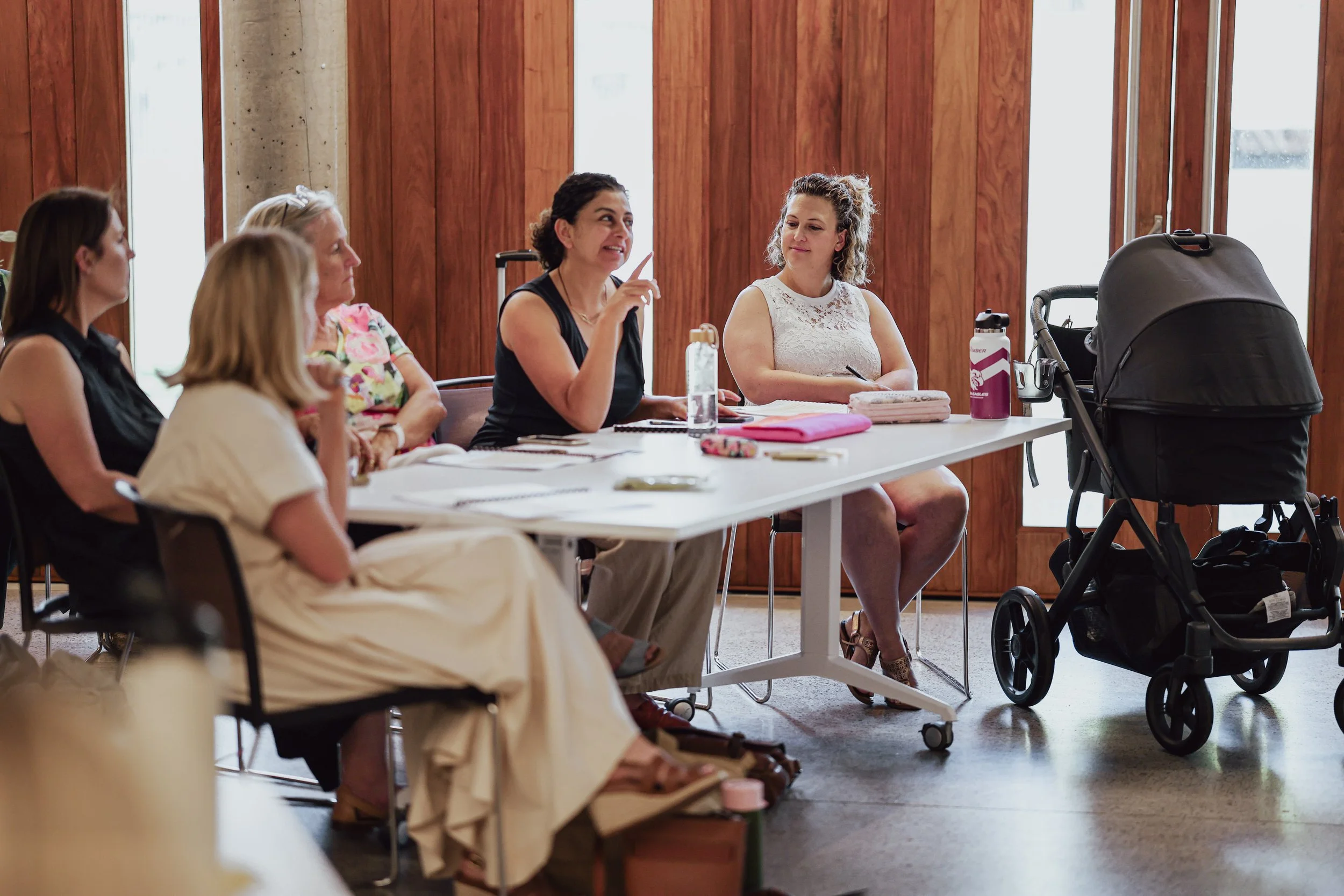 Group of women sitting around a table in a meeting room with wooden walls. One woman is speaking and gesturing with her finger, others are listening. Children’s belongings and a stroller are on the table.