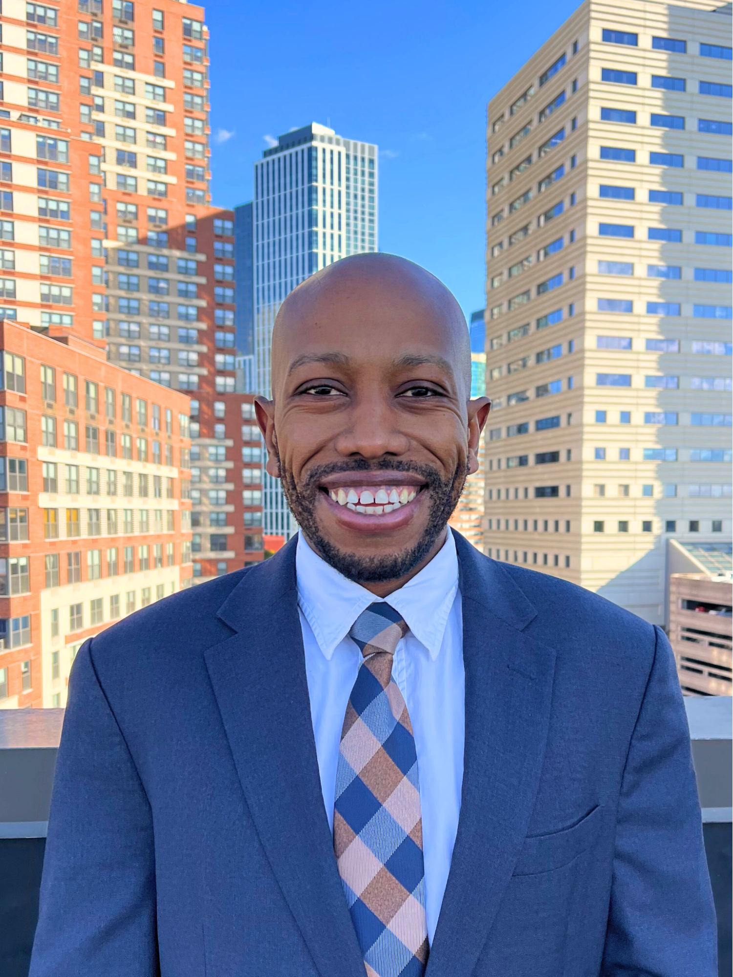 A smiling man in a dark suit, white shirt, and plaid tie standing against a city skyline with tall buildings and a blue sky.