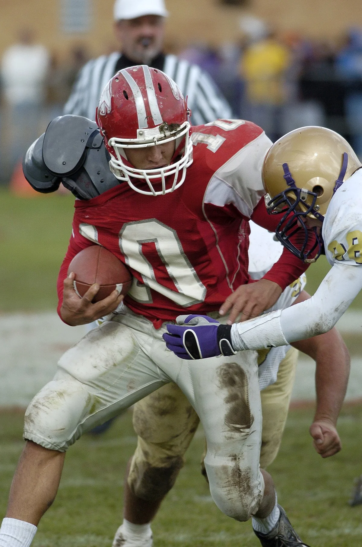 A football player in red and white uniform holding the ball tries to advance while a player in white and gold jersey attempts to tackle him on a muddy field, with a referee and spectators in the background.