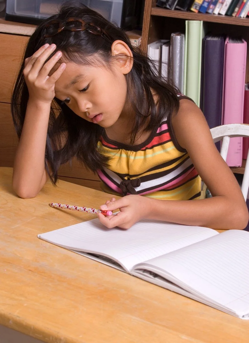 A young girl sitting at a wooden table, holding a pencil, with her head resting on her hand, looking down at an open notebook.