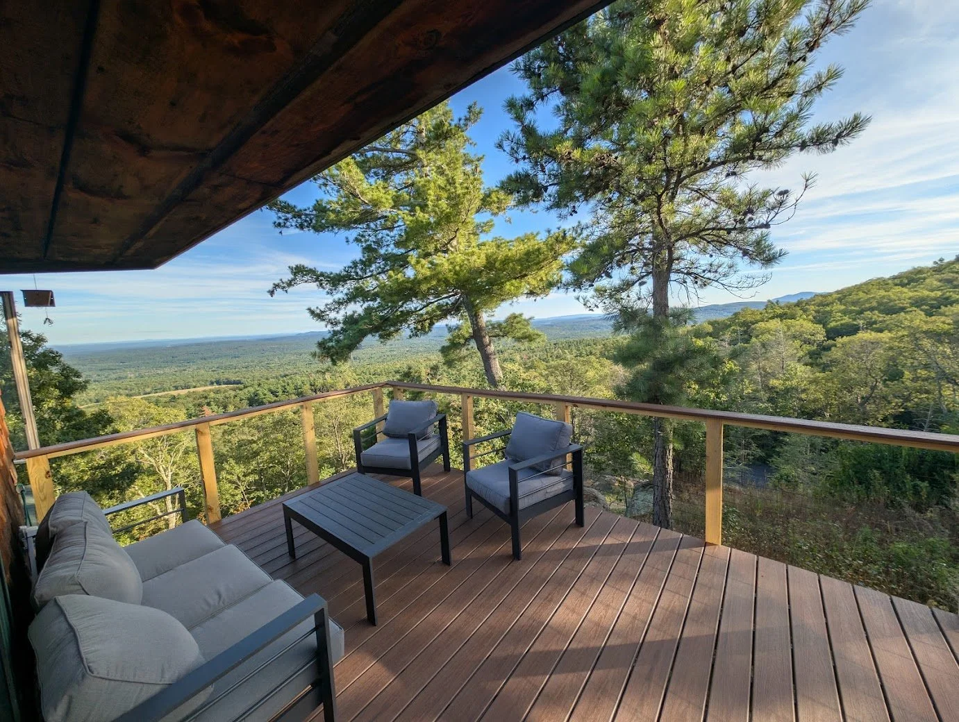 A wooden deck overlooking a forested landscape with tall trees and distant mountains under a blue sky.