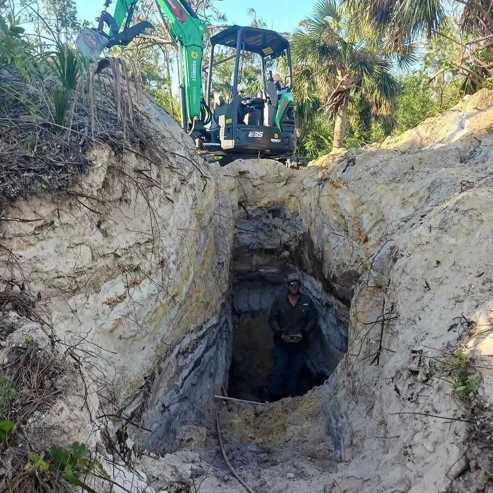 A person standing inside a deep excavated trench with layered rock walls, holding a tool or device, while a mini excavator is parked at the top edge of the trench among vegetation and trees.