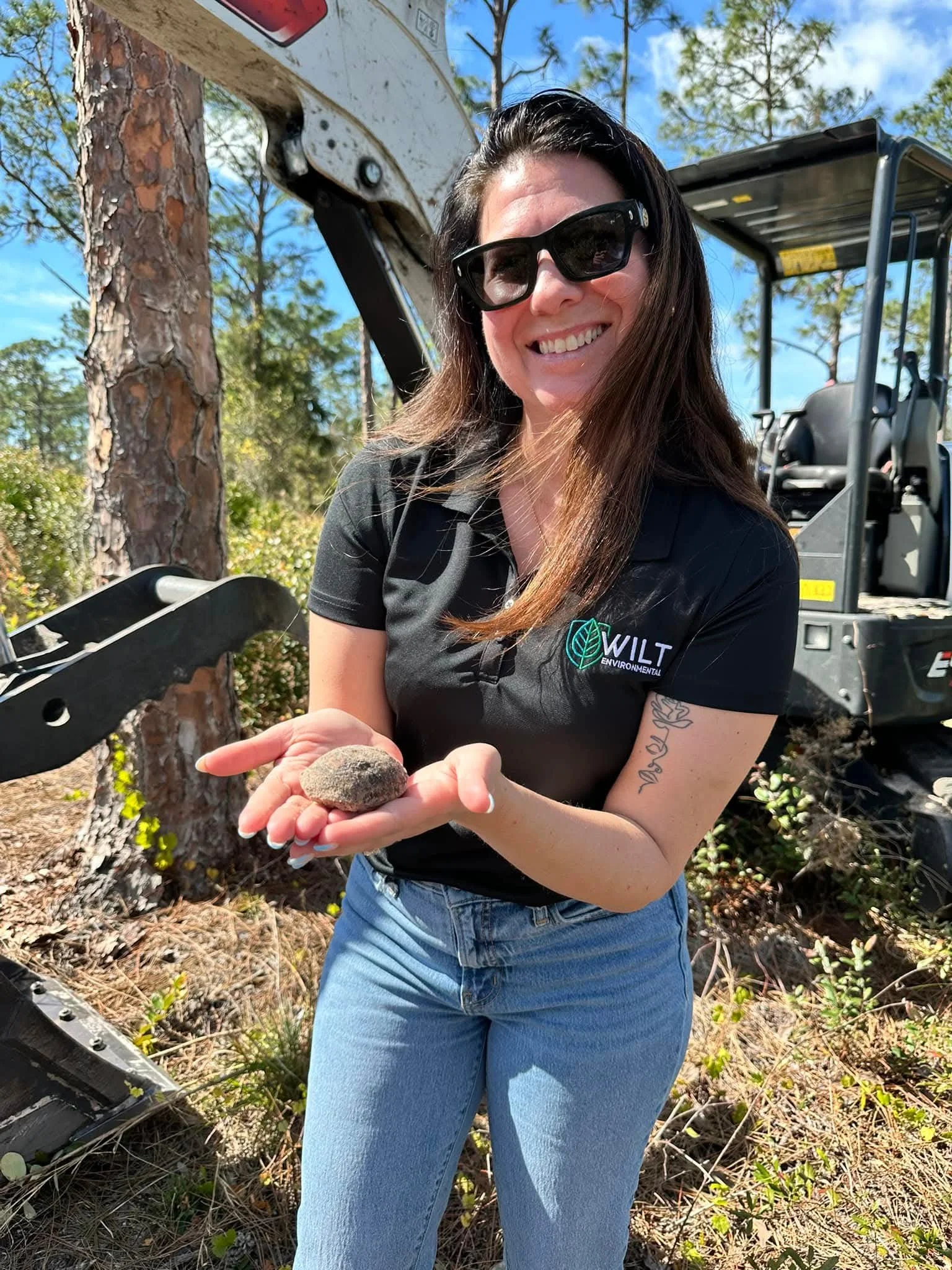 A woman with long brown hair wearing sunglasses and a black polo shirt with 'WILT Environmental' logo, smiling and holding a small rock in her hand while standing outdoors near trees with a construction or forestry vehicle in the background.