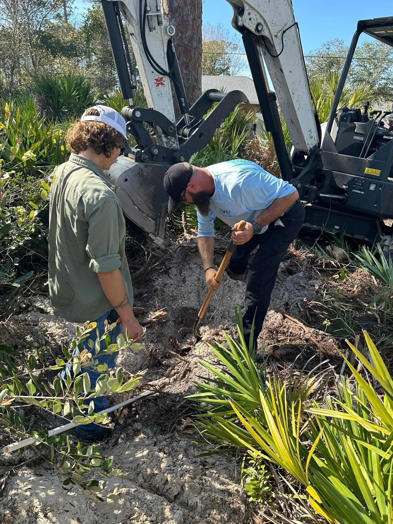 Two men working with a backhoe digging into the ground among green plants and bushes outdoors on a sunny day.