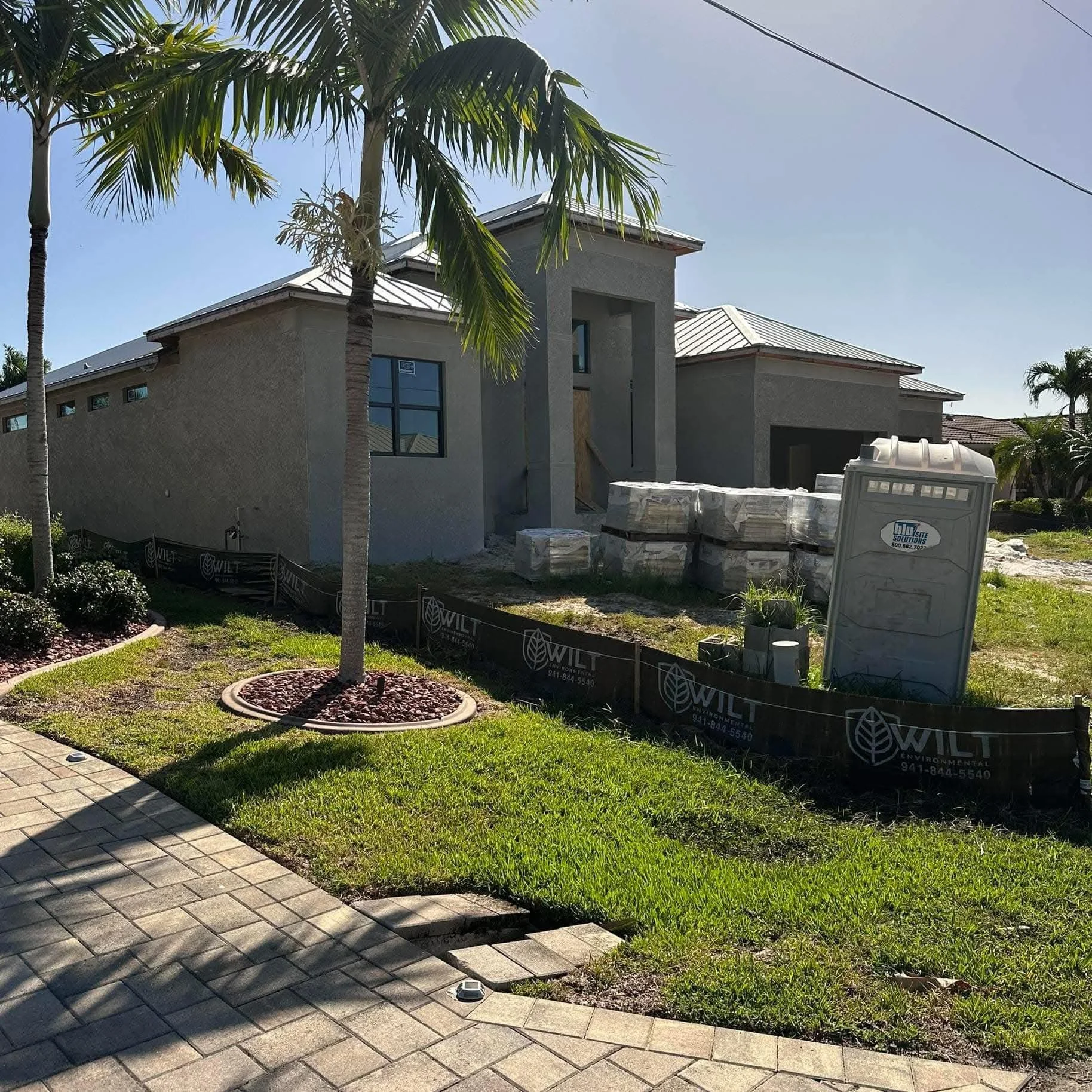 A house under construction with a gray exterior, surrounded by a landscaped yard with palm trees, a paved walkway, and construction materials on the lawn.