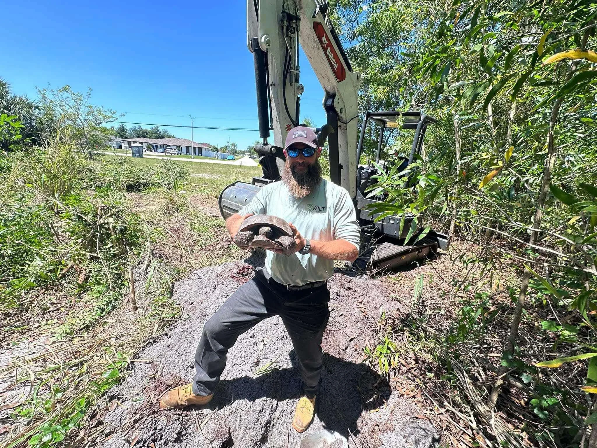 A man with a beard wearing sunglasses, a cap, a light-colored T-shirt, and dark pants stands outdoors on a large fallen tree trunk holding a piece of equipment. Behind him, there is a small excavator in a wooded area with green foliage, and in the ba