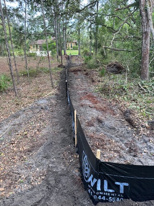 A dirt construction site in a wooded area with a black construction barrier running along the middle and trees on both sides, leading to houses in the background.