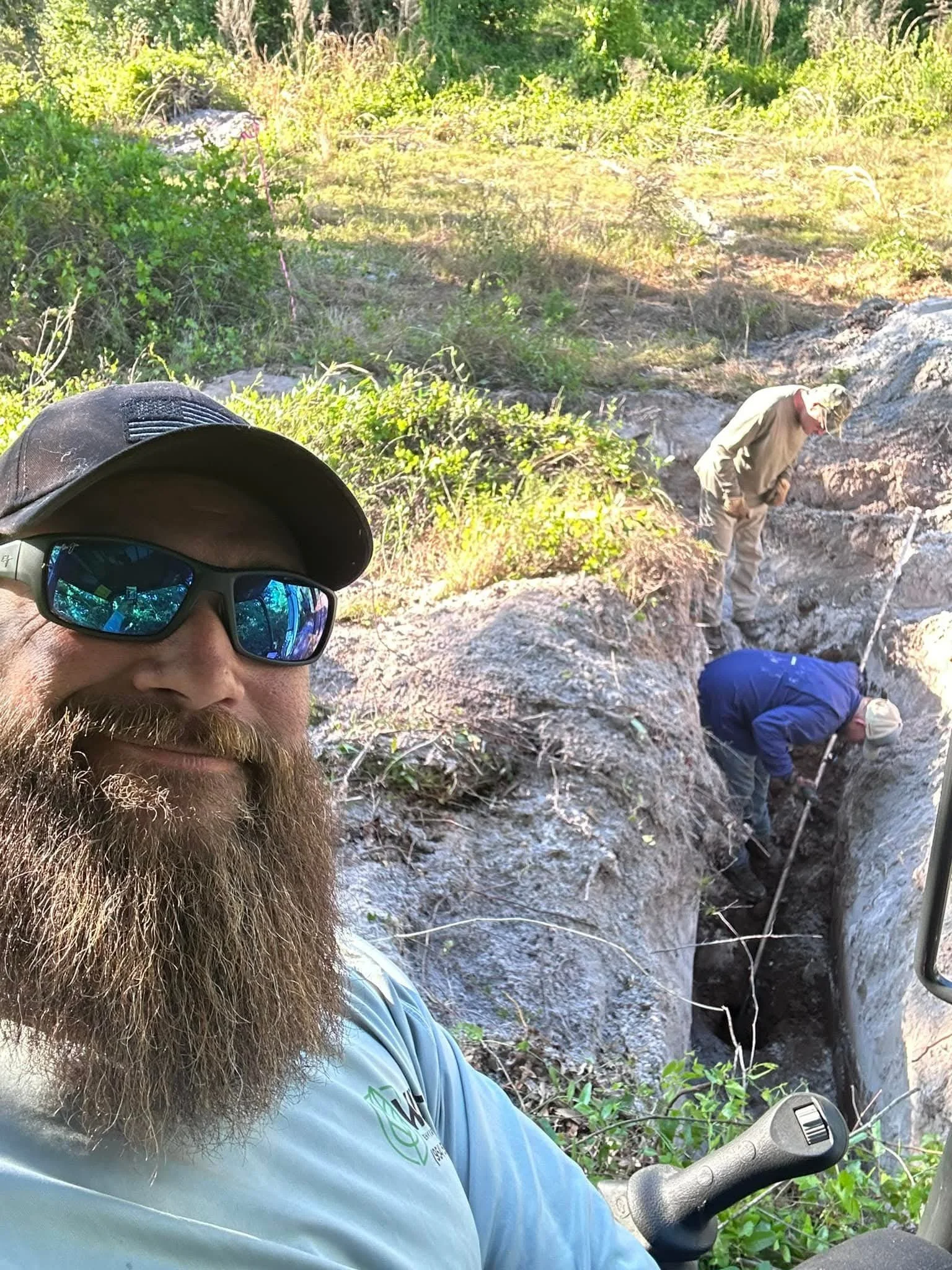 A man with a beard and sunglasses taking a selfie in front of a trench with two other workers digging inside it, surrounded by greenery and rocks.