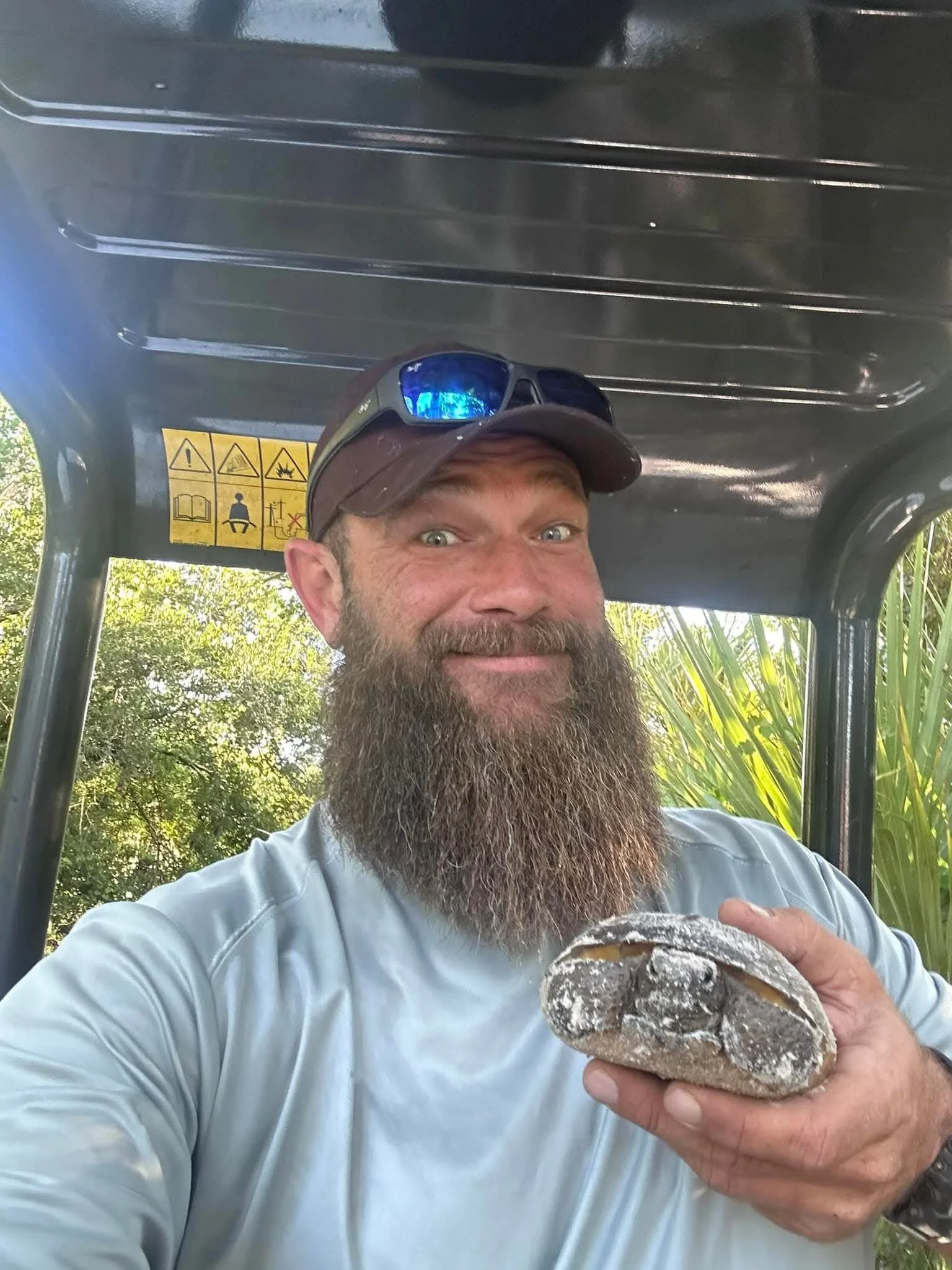 Smiling man with a full beard and sunglasses on his head, sitting in an outdoor cart or vehicle, holding a large rock covered in dust or chalk, with trees and greenery in the background.