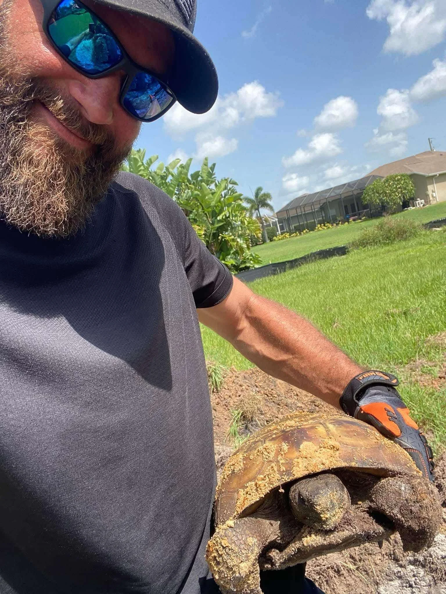 A man with sunglasses and a beard holding a large, weathered turtle shell outdoors on sunny day with houses and greenery in the background.