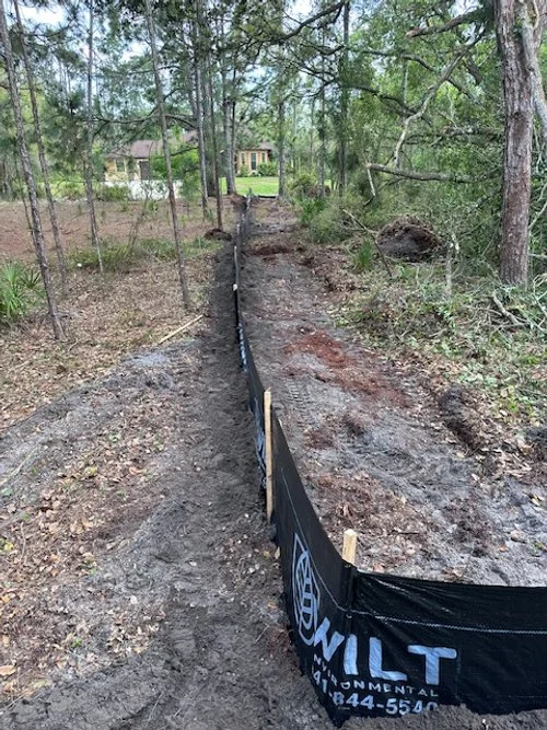 A construction site in a wooded area with a narrow dirt path and a black fabric barrier, possibly for land surveying or excavating work.