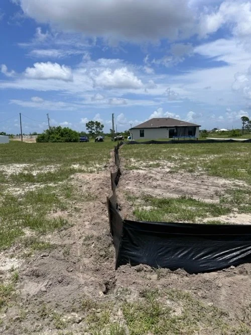 A construction site with a trench dug into the ground, covered partially with black plastic sheeting, near a residential house in a grassy area under a partly cloudy sky.