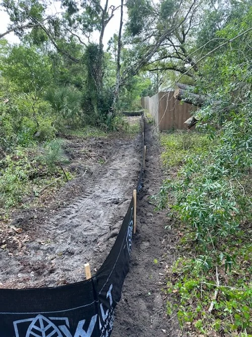 A cleared dirt path with a black construction fence alongside, surrounded by green trees and bushes, and a wooden fence in the background.