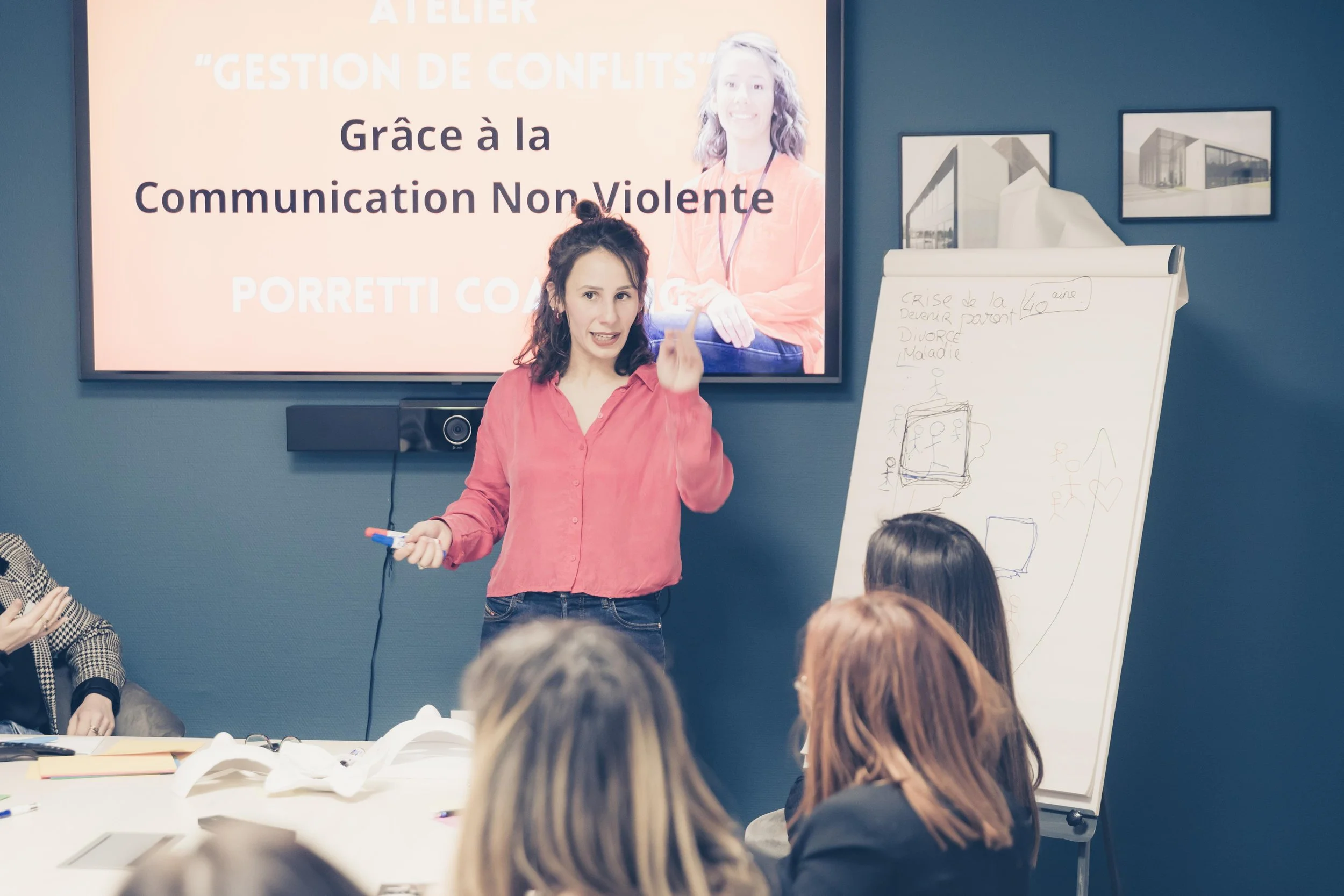 Une femme en train de donner une présentation à un groupe de personnes dans une salle de formation. Un grand écran derrière elle affiche un message en français sur la gestion de conflits et la communication non violente.