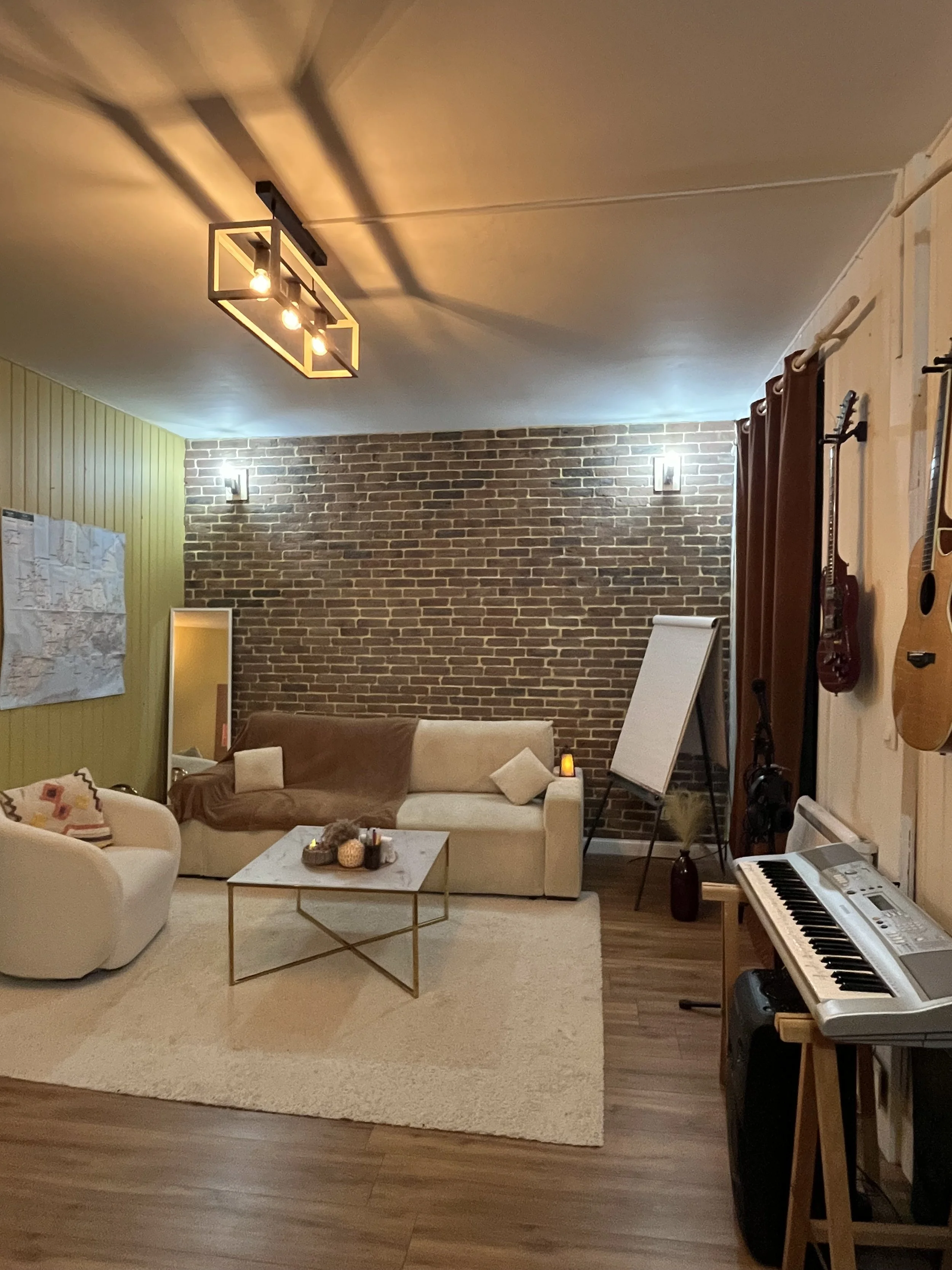 Salon de musique et de détente avec un canapé beige, un tapis clair, un mur en briques, une table basse avec des bougies, une guitare accroché à un mur, un clavier électrique, et une étagère avec une enceinte.