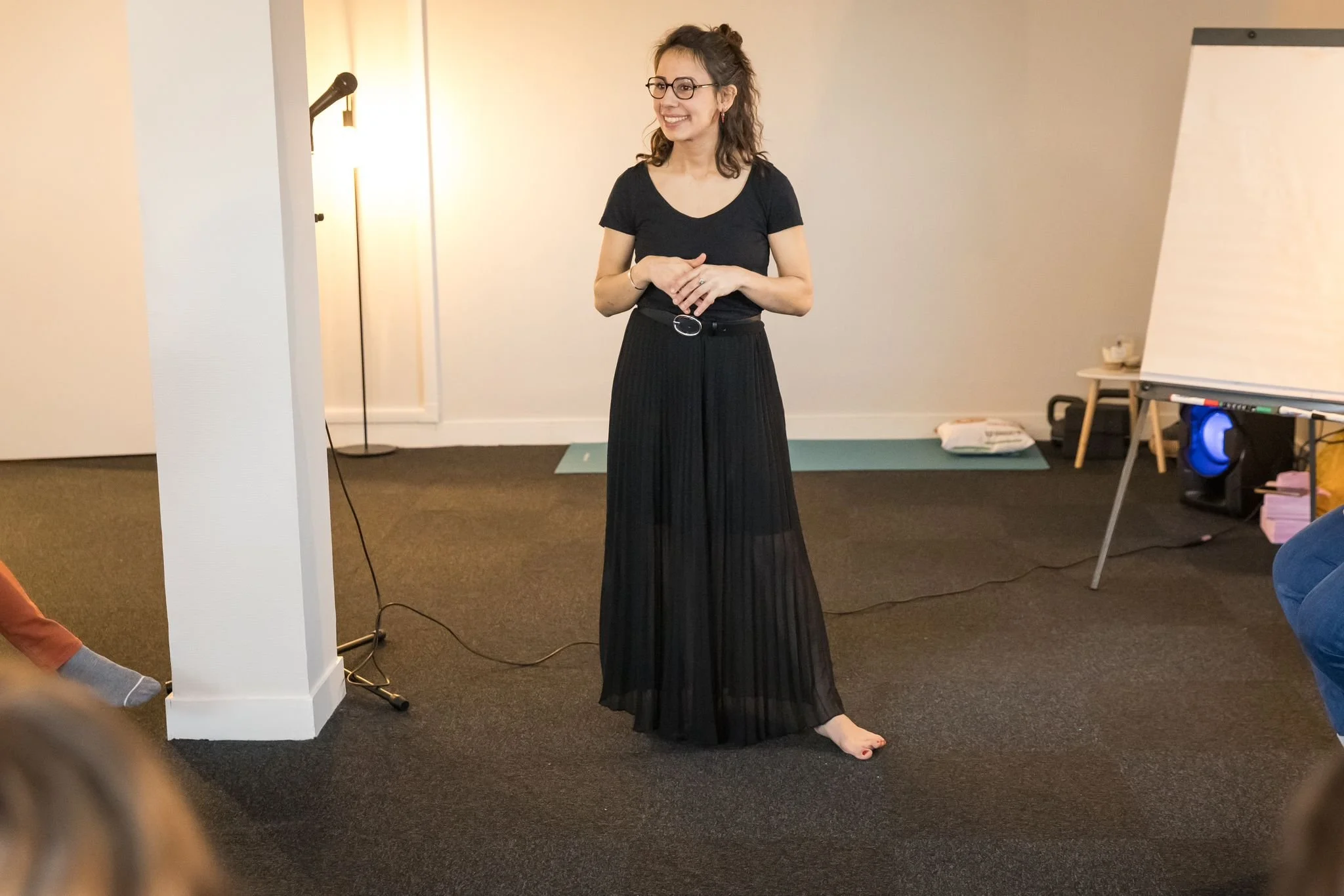 Une femme souriante portant des lunettes, des vêtements noirs, et debout sans chaussures, donne une présentation ou un discours dans une pièce, avec un tableau blanc à sa droite et un lampadaire derrière elle.