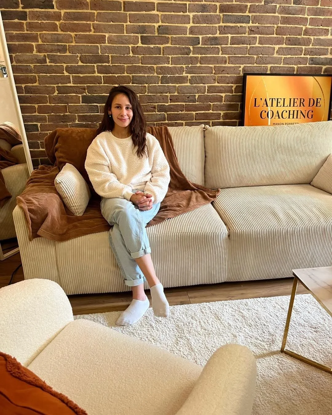 Une femme assise sur un canapé beige rayé, coiffée de cheveux longs et bruns, portant un pull blanc, un pantalon beige et des chaussettes blanches, dans un salon avec un mur en briques et une télévision à l'arrière affichant 'L'ATELIER DE COACHING'.