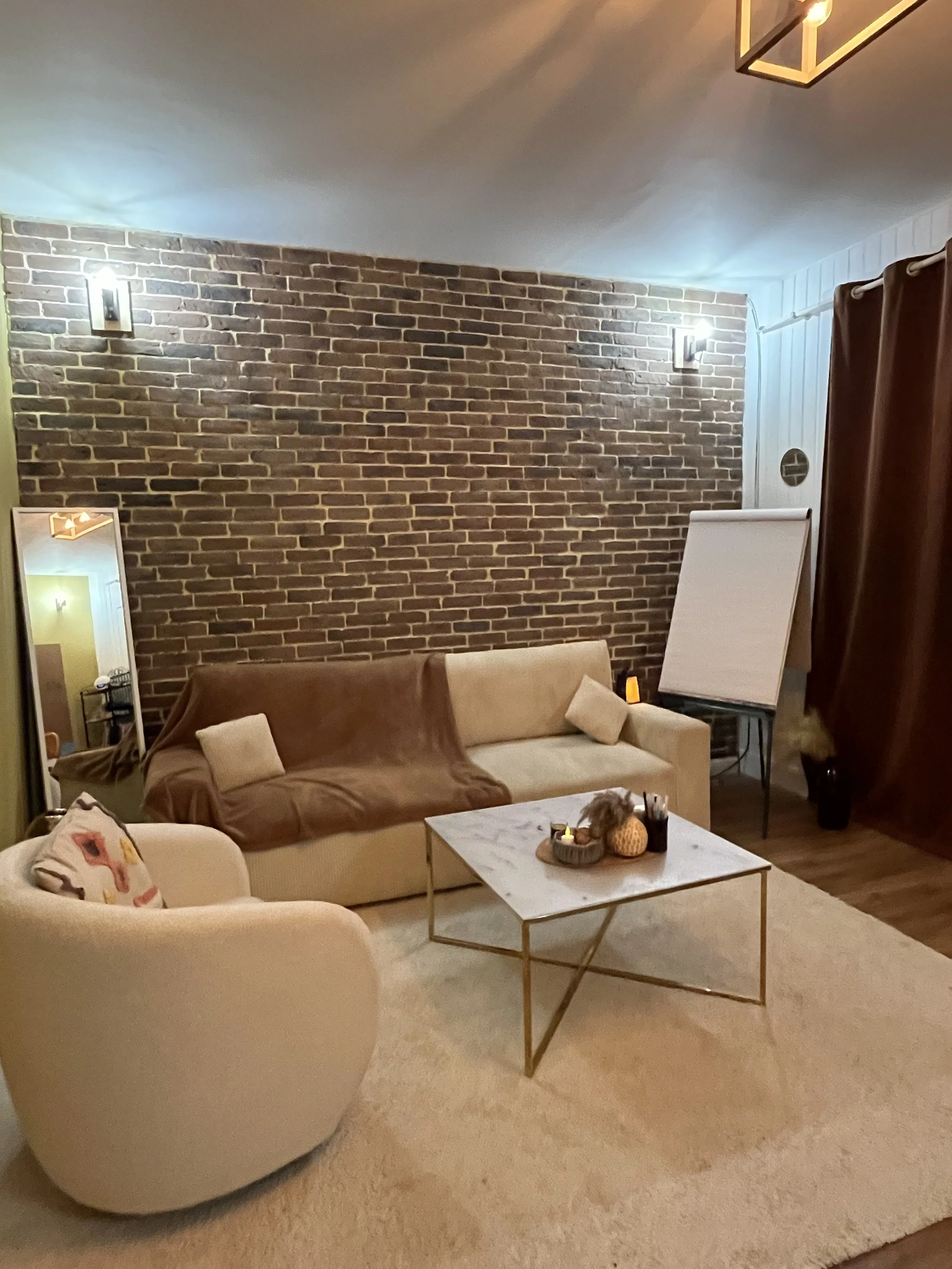 Salon avec canapé beige, fauteuil blanc, table basse en marbre et métal, mur en briques apparentes, miroir, éclairage chaleureux, rideaux marrons et tapis beige.