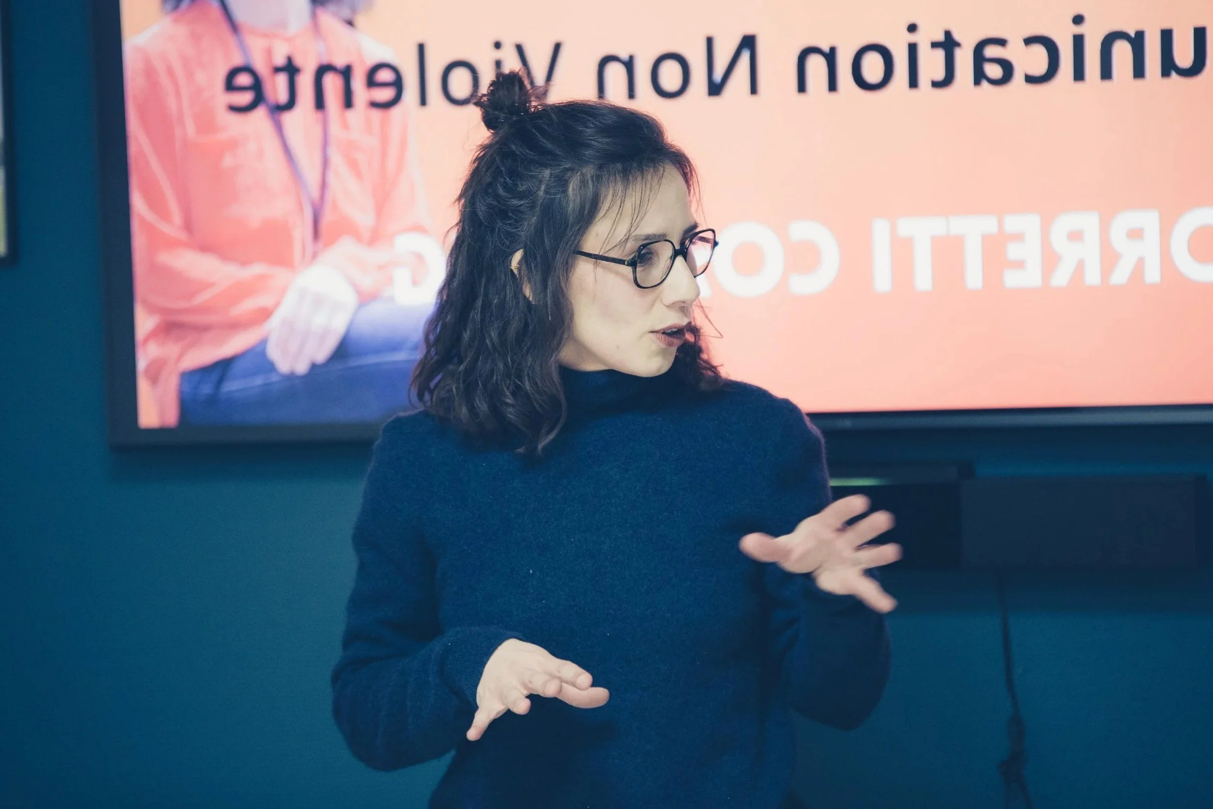 Une femme avec des lunettes noires, portant un pull noir, gesticulant avec ses mains, se trouve devant un écran affichant une image d'une autre personne en arrière-plan.