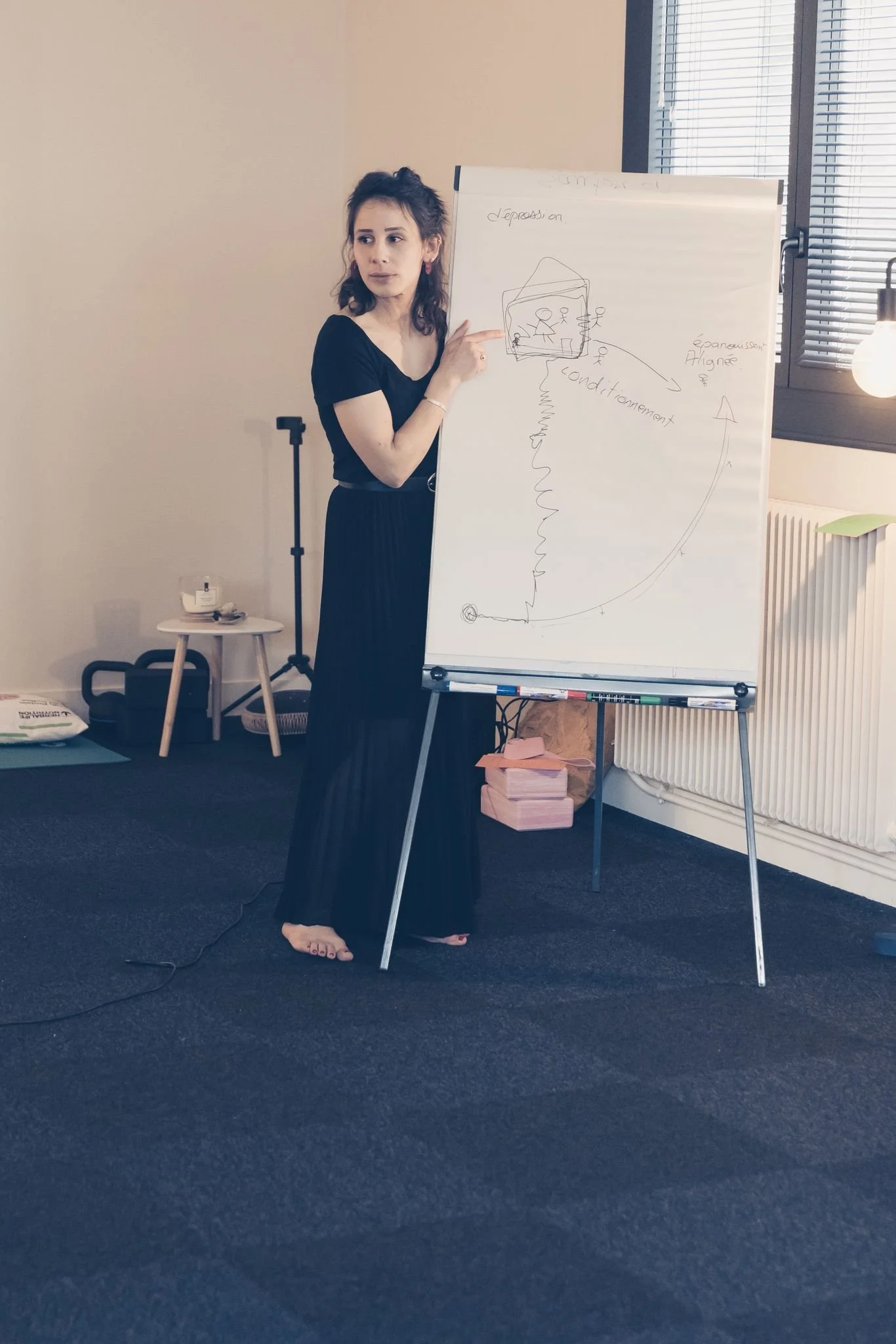 Une femme en vêtements noirs explique un schéma sur un tableau blanc dans une salle de formation ou de réunion.