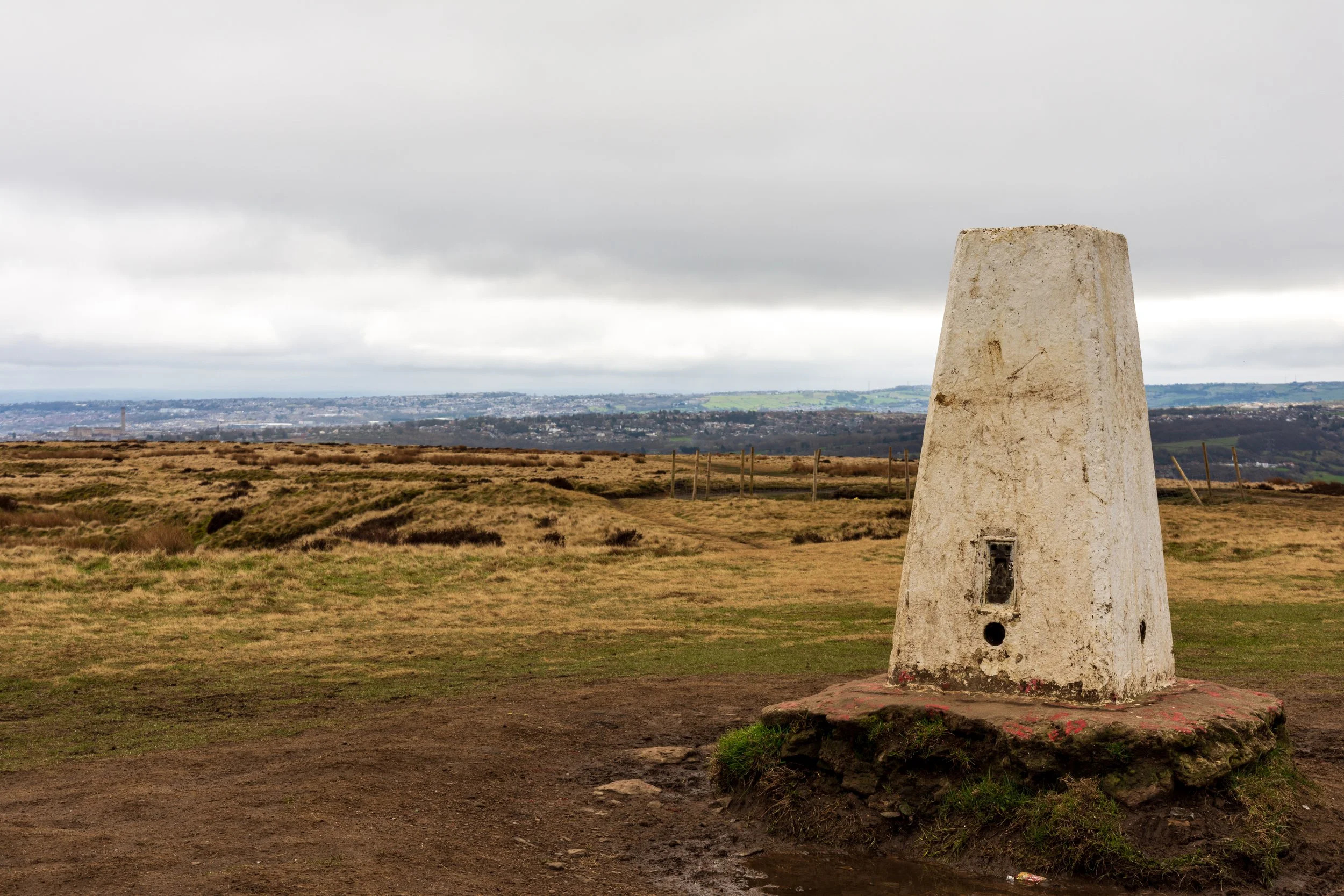 A trig point on the summit of Baildon Hill, with a view over Bradford, under a cloudy sky
