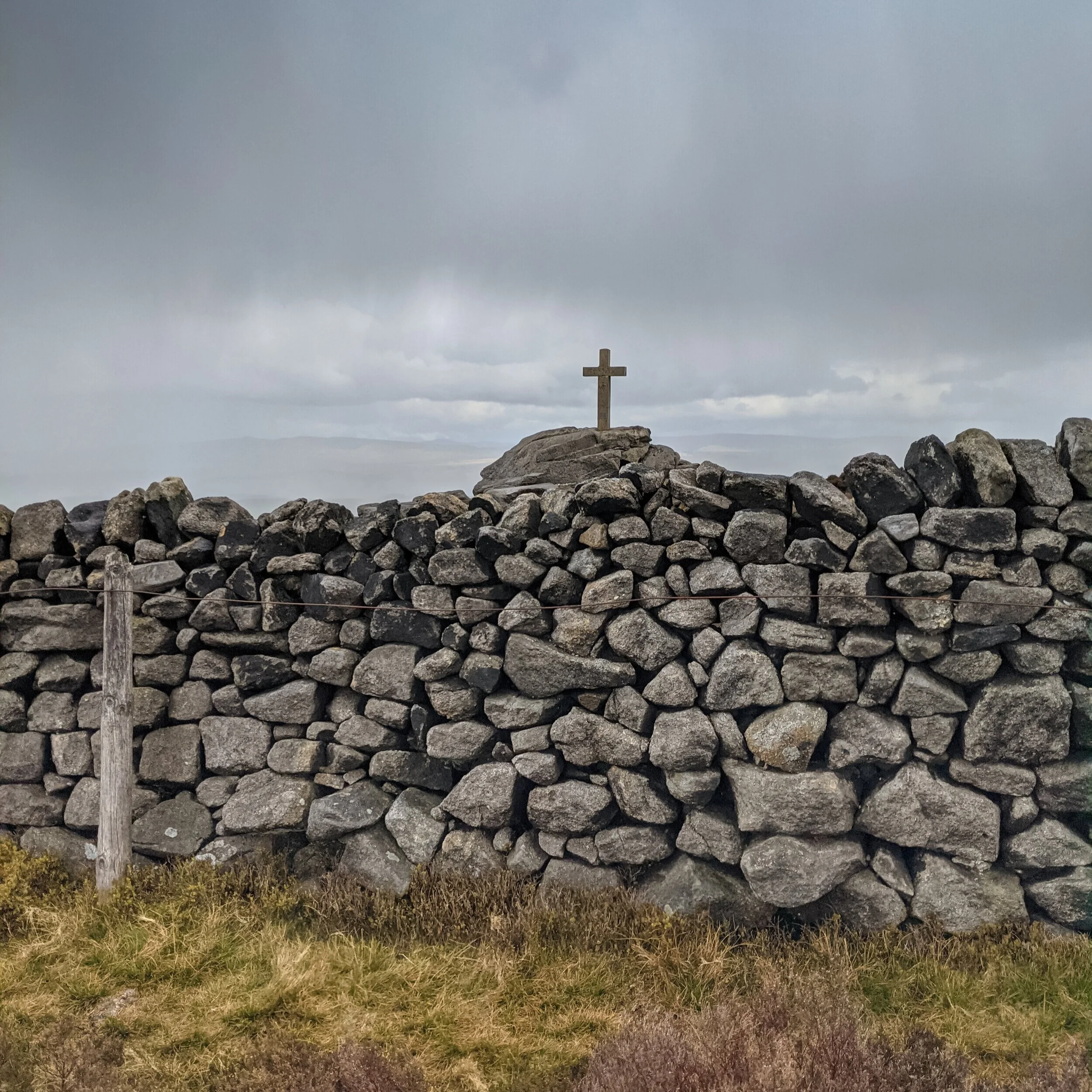 The now-concrete Rylstone Cross on Cracoe Fell, with a drystone wall in the foreground, and a rainy backdrop across the Yorkshire Dales